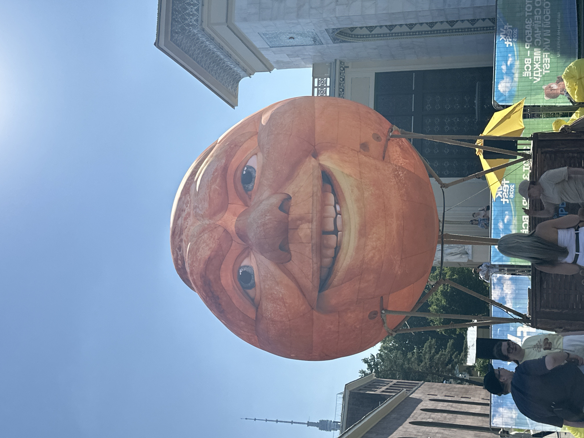 The image shows a large, orange, inflatable balloon in the shape of a human face with a wide smile, positioned outdoors under a clear blue sky. The balloon is supported by a wooden structure. In the background, there are buildings with marble facades and decorative elements, as well as trees and a tall communication tower. Several people are visible in the foreground and middle ground, some looking at the balloon, others engaged in conversation. There is also a yellow umbrella and a banner with Cyrillic text. This scene is located at VDNKh in Moscow, Russia, specifically near the Cosmos Pavilion.