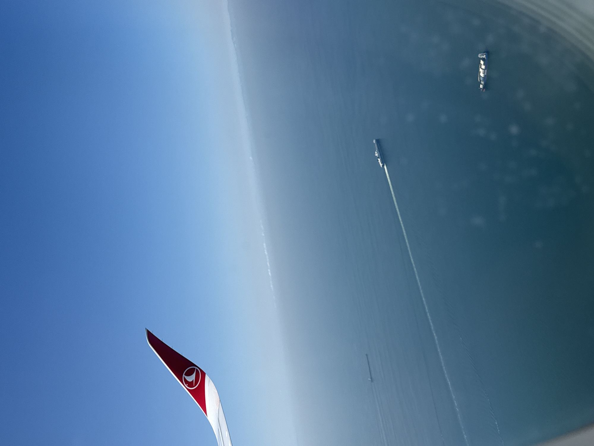 An aerial view from an airplane shows a clear blue sky above a calm, deep blue sea. The wing of the airplane, adorned with the Turkish Airlines logo, is visible in the upper left quadrant of the image. Several ships are scattered across the water. One large vessel is in the distance on the left, a long ship is moving diagonally across the mid-section of the image, leaving a white wake, and a smaller ship, likely a container ship, is in the lower right. The horizon is visible in the distance, with a faint haze obscuring landmass.
