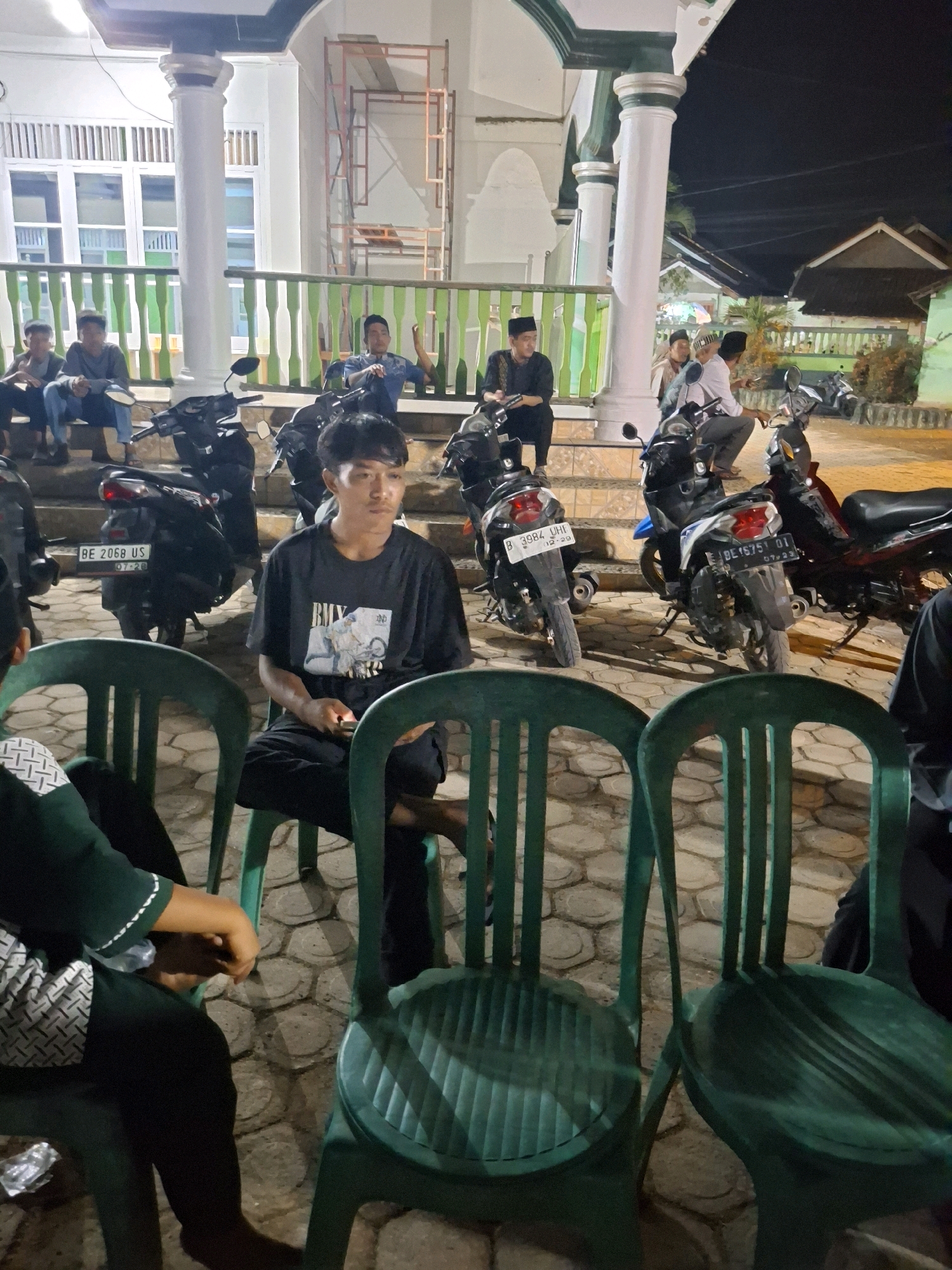 The image depicts an outdoor scene at night, likely in front of a mosque or similar building in Yogyakarta, Indonesia. Several people are visible, some sitting and others standing. In the foreground, two green plastic chairs are prominently featured. A young man is seated in one of the chairs, wearing a black t-shirt with a graphic print and black pants. He appears to be holding a small object. Another person, wearing a dark green shirt and black pants, is seated in the other chair, with their hands resting on their lap. Behind them, several motorcycles are parked. In the background, a group of men, some wearing traditional Islamic attire and caps, are gathered near the building entrance. The building itself has white pillars with green accents and a patterned white facade. Scaffolding is visible on one side of the building, suggesting ongoing construction or renovation. The ground is paved with hexagonal stones. The overall atmosphere appears relaxed, with people gathered casually.