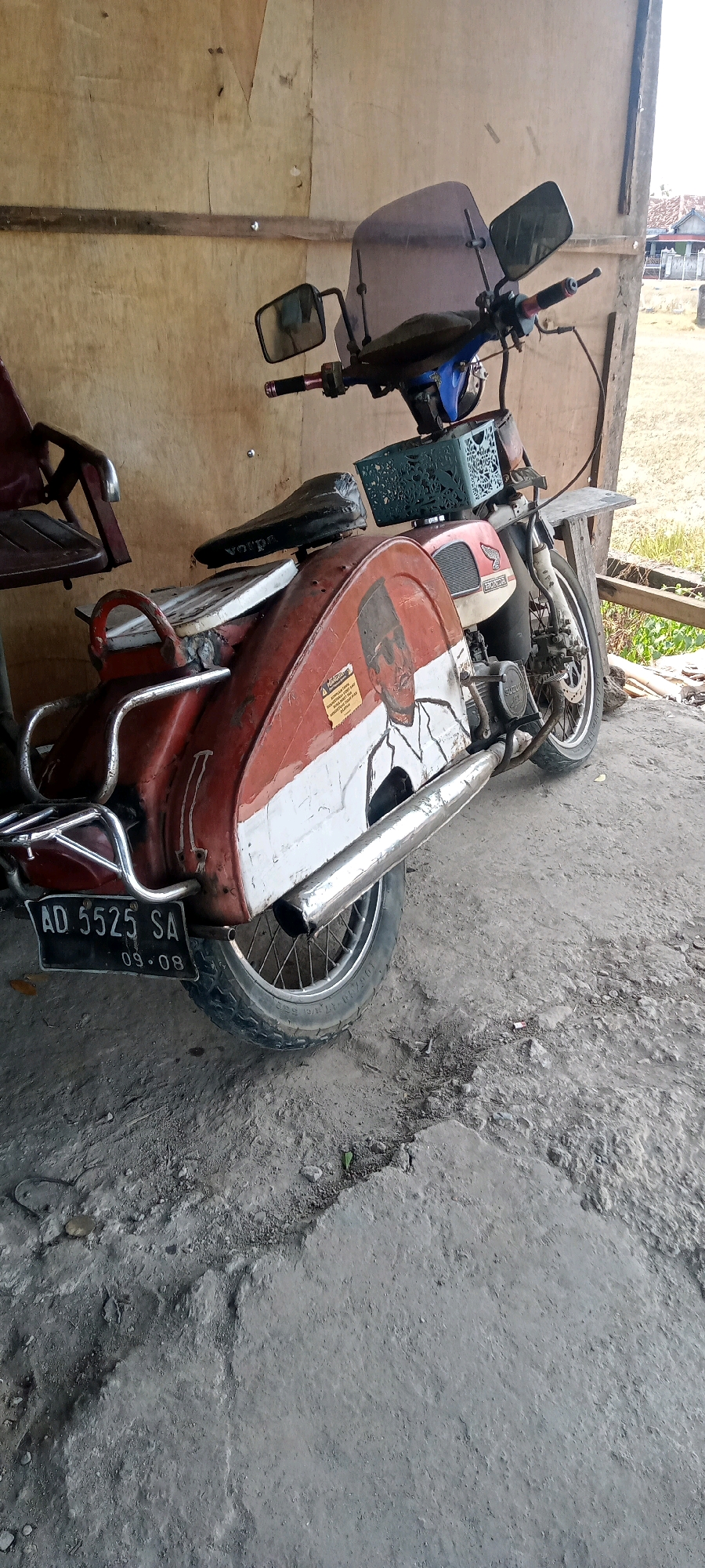 The image shows a vintage Honda motorcycle parked on a concrete floor under a wooden structure in the city of Bulu, Indonesia. The motorcycle is red and white with a painting of a man in traditional Indonesian attire on the rear fender. A license plate reading "AD 5525 SA 09-08" is attached to the back. The environment appears to be an outdoor area, possibly a workshop or storage space, with a wooden wall behind the motorcycle and dry grass and buildings visible in the background. The scene is well-lit by natural light.