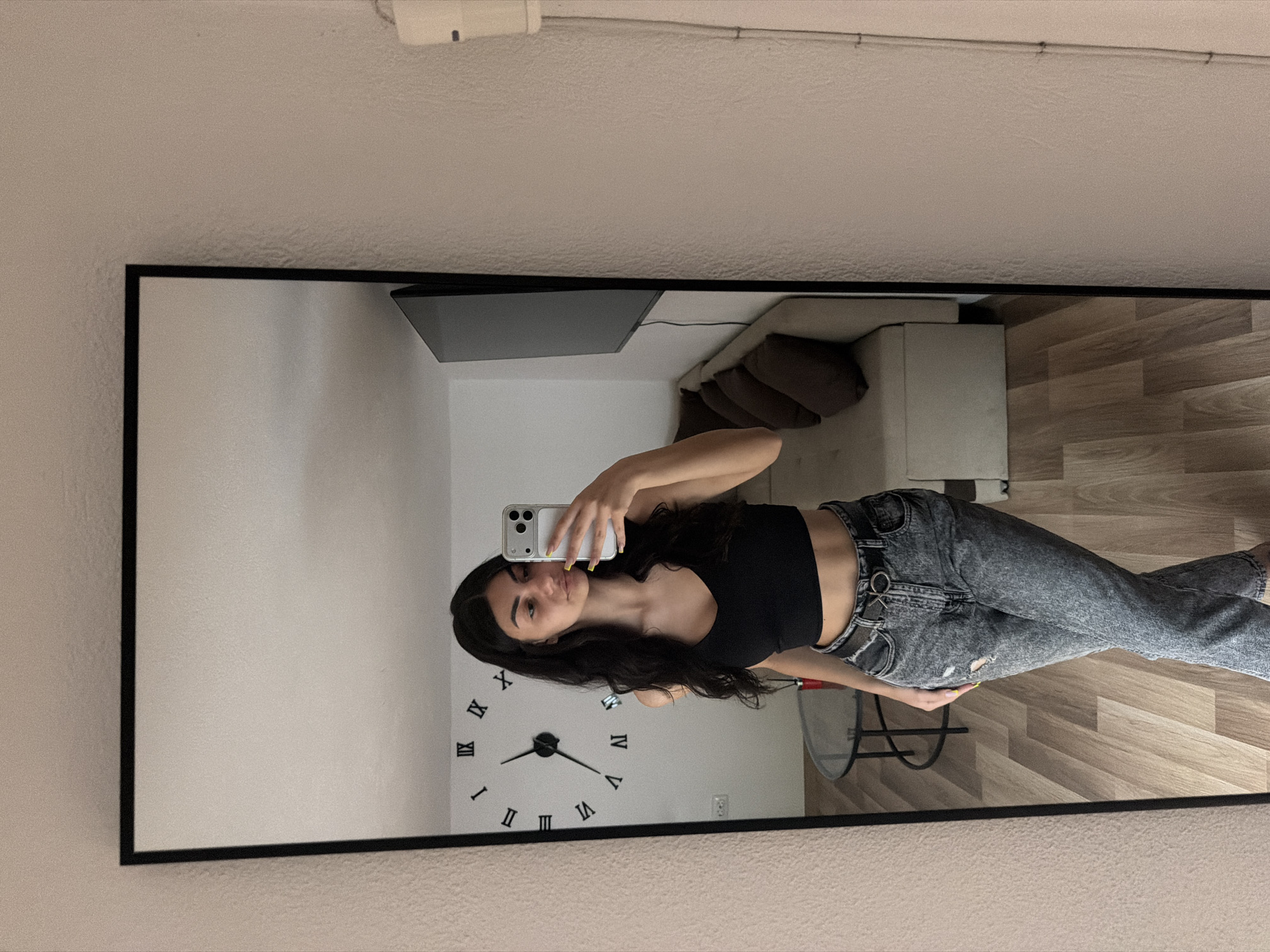 A young woman captures a mirror selfie in an indoor setting in Gabrovo, Bulgaria. She is positioned slightly to the left within the frame of a large, black-bordered rectangular mirror, holding a white smartphone with multiple cameras in her reflected left hand (her right hand). She has long, dark hair and is wearing a black crop top, exposing her midriff, and acid-wash gray ripped jeans with a belt featuring a silver O-ring buckle. Her fingernails are painted yellow.
The reflection reveals a light-colored, textured wall behind her, adorned with a prominent wall clock featuring black Roman numerals (I through XII) and hands indicating approximately 5:50. To her right, a light-colored sofa with brown cushions and a dark-colored television or monitor mounted on the wall are visible. The room also features a light-toned wooden floor. The ceiling shows some visible wiring or a fixture. The woman maintains a neutral to slightly pouty expression, typical for a selfie pose. The overall setting appears to be a modern and personal living space.