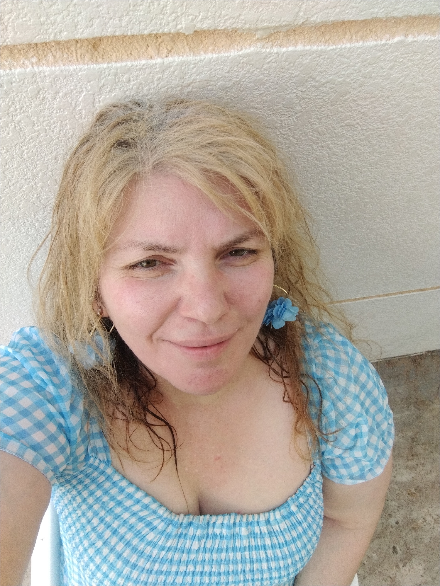 A woman is centered in the frame, likely in Al Mas'oudi, United Arab Emirates, based on the request. She is fair-skinned, with blonde, shoulder-length hair that appears to be partially wet. Her hair shows gray at the roots. She has hazel eyes, and she is smiling subtly.
She is wearing a blue and white gingham top with a low-cut neckline. A blue flower earring is visible on her right side, and a gold hoop earring is visible on her left. The background is a textured, off-white wall. The bottom of the frame shows a pale concrete surface.