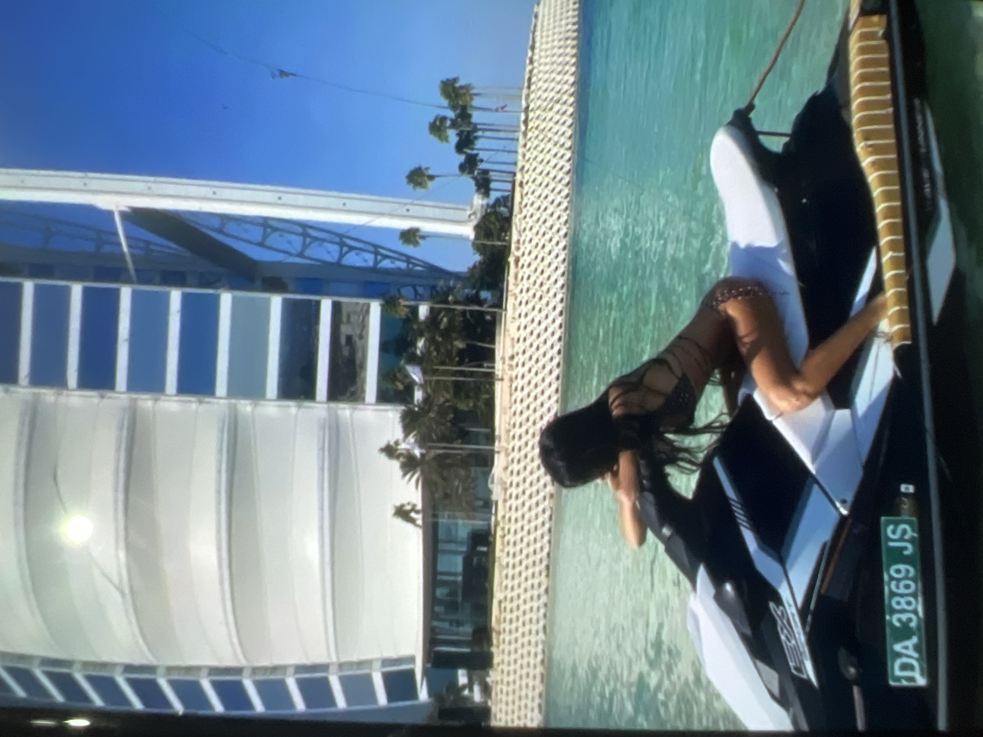 A woman with dark hair, wearing a patterned bikini, is seated on a black and white jet ski on a bright turquoise body of water. She is leaning forward, resting her forearms on the handlebars. Her legs are bent, and her right knee is visible. The jet ski has a green license plate in the foreground displaying "DA 3869 J$". In the background, a modern, sail-shaped building with a white exterior and blue-tinted windows rises against a clear blue sky. Palm trees and other vegetation are visible in front of the building, along with a textured, light-colored wall made of interlocking blocks. The scene appears to be a sunny day, with clear water and a vast sky.