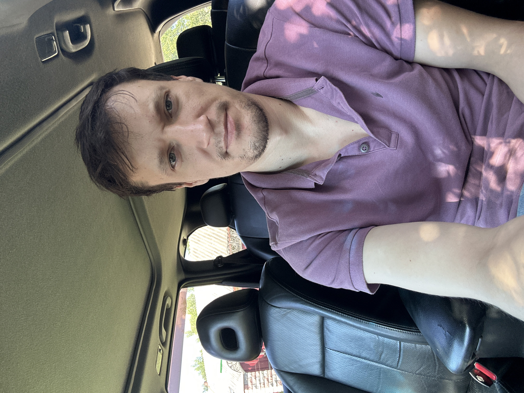 A selfie is taken from inside a car, showing a man in a purple polo shirt sitting in the driver's seat. The man has short brown hair and a light beard. The interior of the car is visible, including black leather seats, a sunroof, and the car's ceiling. Outside the car, through the windows, a brick building and trees can be seen. The car is parked outdoors on a sunny day. The location is identified as Shatava, Ukraine.