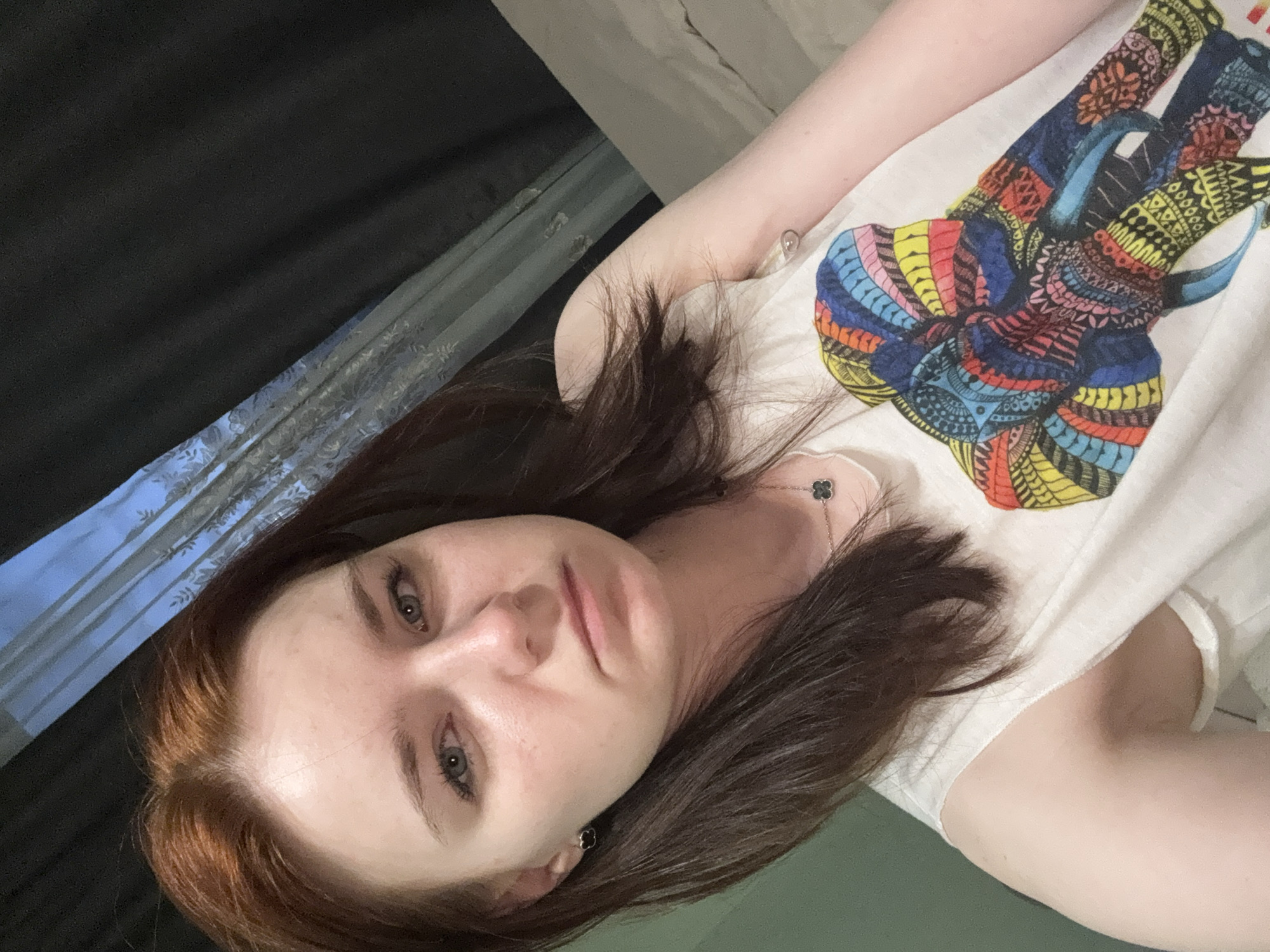 A woman with brown hair and pale skin is lying down, looking at the camera. She is wearing a white t-shirt with a colorful, stylized elephant graphic. Behind her are dark curtains and a glimpse of a window. The location is Unknown, Unknown.