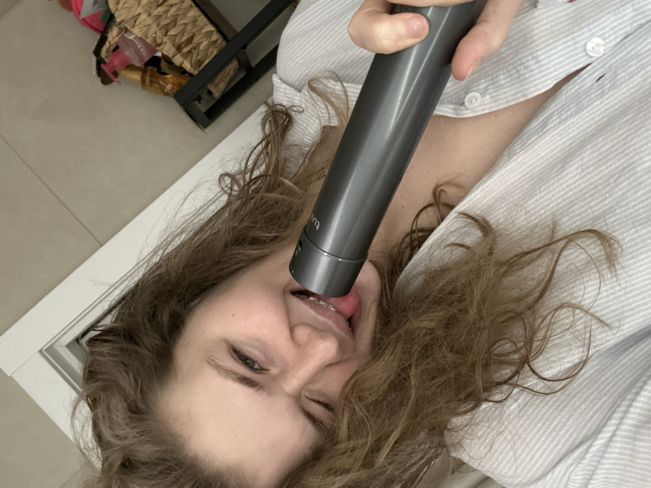 A woman with braces lies on her back, winking at the camera. She wears a striped shirt and has curly brown hair. A cylindrical gray object is held in front of her face, with one end near her open mouth and tongue. A portion of a doorway is visible in the lower left corner, and a shelf containing various items is in the upper left. The floor tiles can be seen in the background.