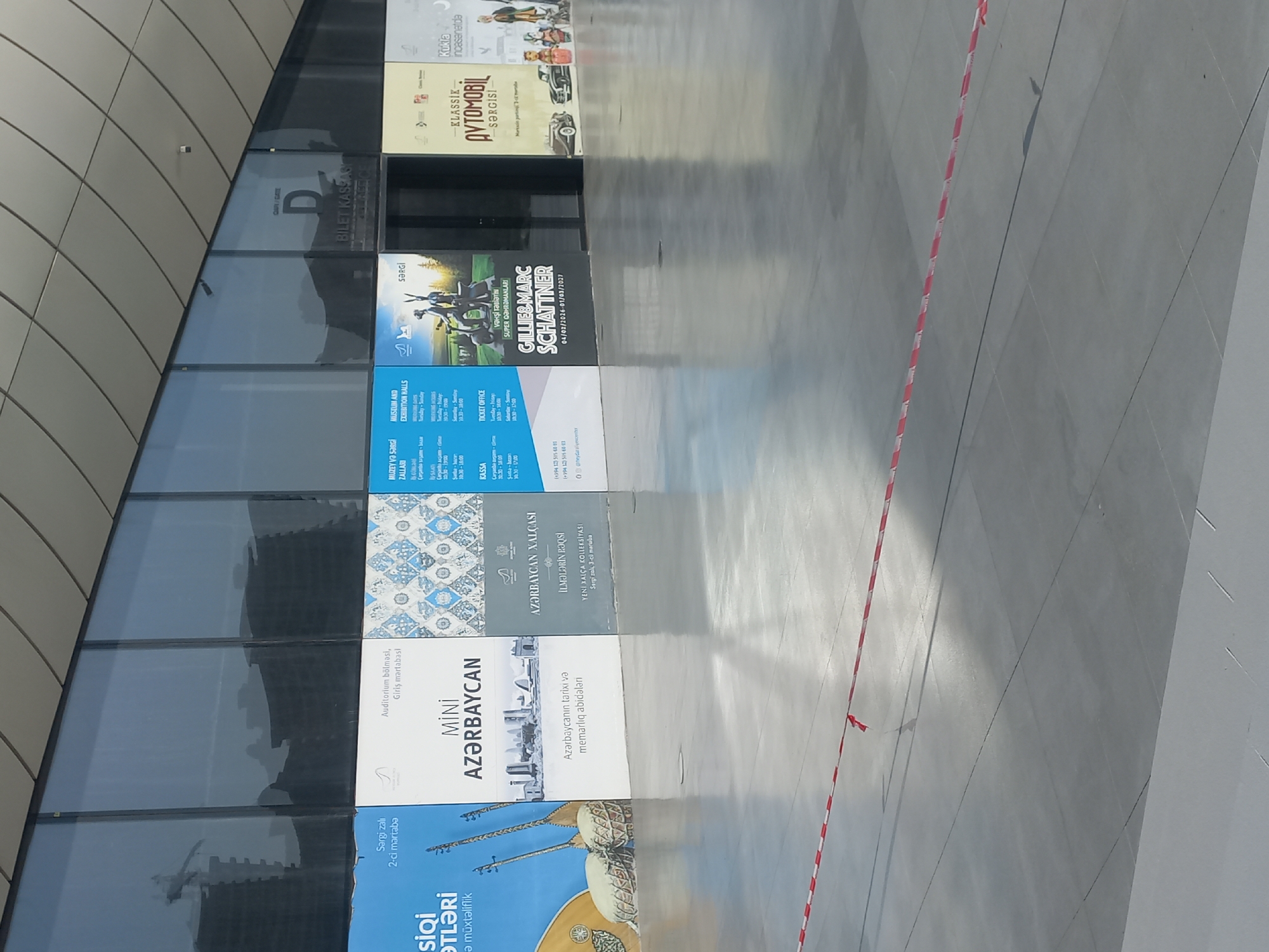 The image shows a wide shot of a modern building's exterior, likely an exhibition center or museum, in Baku, Azerbaijan. The building features a sleek, dark-colored facade with large glass panels reflecting the sky and surrounding structures. In front of the building, several large banners or posters are displayed, advertising various exhibitions and events.
From left to right, the posters include:
- One with the title "SİQİ ƏTLƏRİ" in large font, with the subtitle "Ən müxtəliflik" below. This poster features imagery of musical instruments.
- A poster for "MİNİ AZƏRBAYCAN," which appears to showcase historical sites or miniatures of Azerbaijan.
- A poster with "AZƏRBAYCAN XALÇASI" and "İLMƏLƏRİN RAQSI" and "YENİ XALÇA KOLLEKSİYASİ," suggesting an exhibition on Azerbaijani carpets.
- A poster with "MUSEUM AND EXHIBITION HALLS" and "BILET KASSASI" (Ticket Office), indicating functional aspects of the venue.
- A poster for an exhibition titled "GILLIE & MARC SCHATTNER" with dates "04/02/2026 - 01/03/2027."
- A poster for "KLASSİK AVTOMOBİL SƏRGİSİ," an exhibition of classic automobiles.
- A poster for "Kukla inçasenetda," which likely refers to puppet art or theater.
The foreground is a smooth, grey concrete floor, marked with faint grid lines. A red and white caution tape is stretched across the floor in a diagonal line, suggesting a restricted or cordoned-off area. The overall atmosphere is clean and modern, characteristic of contemporary public spaces in a major city. The architectural style and the presence of specific exhibition themes point towards a cultural venue in Baku.