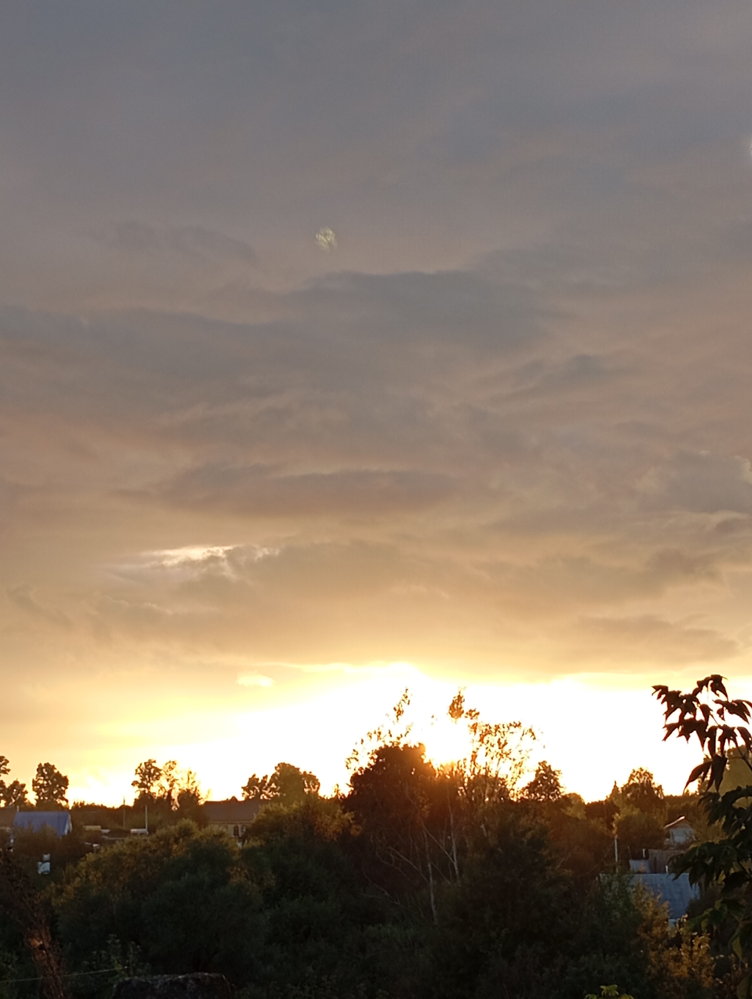 The image depicts a sunset scene over a rural landscape, likely in the city of Fokino, Russia. The sky is filled with a mix of grey and orange clouds, with a bright, sunlit area near the horizon. A faint, circular, luminous object is visible in the upper part of the sky, possibly a lens flare or an atmospheric phenomenon. Below the sky, a dense line of trees and vegetation forms the midground. Several houses with blue and grey roofs are partially visible through the trees in the distance. The foreground shows dark foliage and a rough, dark surface, possibly a wall or ground. The overall impression is of a tranquil evening in a rural setting.