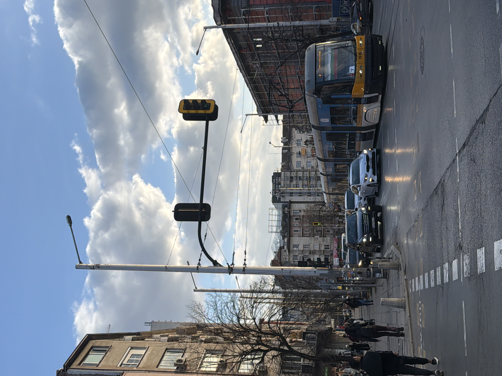 This image depicts a bustling urban street scene in Sofia, Bulgaria, characterized by vehicular and pedestrian traffic under a partly cloudy sky. The pavement is wet, suggesting recent precipitation.
In the foreground on the left, two pedestrians walk away from the camera on a wet sidewalk. The closer individual is male, wearing a dark jacket and trousers with white sneakers. The person behind them is female, wearing a dark jacket and trousers. White rectangular pedestrian crossing markings are visible on the wet asphalt road.
In the midground, a modern blue, yellow, and white tram, displaying "7" and vehicle number "2315" along with a Bulgarian flag on its front, moves towards the viewer on the right side of the street. Adjacent to it, a black Mercedes-Benz SUV with Bulgarian license plate P.K 4366 TT travels away from the camera. Behind the SUV, a white sedan with Bulgarian license plate CB 1200 HE follows. Overhead, a black traffic light displays a green signal. Multiple utility poles and overhead tram lines crisscross the scene. Bare deciduous trees line the left sidewalk.
On the left side of the street, a multi-story building with a worn, light-colored facade, numerous windows, and external air conditioning units rises. On the right, a large building is undergoing renovation, evidenced by extensive scaffolding covering its reddish-brown exterior. Further in the background, additional multi-story urban buildings are visible, including one partially identifying "HOTEL CENTRAL PLAZA."
The background reveals a distant snow-capped mountain range under a sky filled with a mix of blue patches and large, white cumulus clouds.
