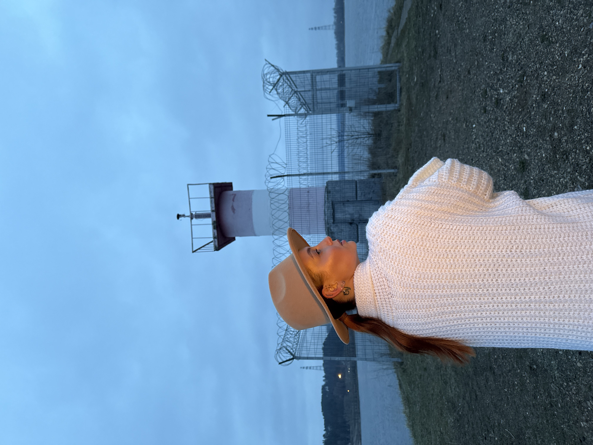 This image, presented as if correctly oriented, captures an outdoor scene in Chaikovsky, Russia, depicting a person by a body of water under an overcast sky. In the foreground, on the right, a person is shown in profile, looking upward and slightly to the left with a contemplative expression. They are wearing a light-colored, possibly off-white, chunky knit sweater and a light brown fedora-style hat. Long, reddish-brown hair cascades from behind their shoulder. Small, ornate earrings are visible on their left ear, along with what appears to be a small tattoo near the earlobe.
In the mid-ground, a red and white striped lighthouse stands on the left, acting as a navigational aid. A chain-link fence topped with razor wire runs across the scene, extending from the foreground towards the distant background, separating the land from the body of water. This water body, likely a river or reservoir, stretches across the middle ground, reflecting the muted blue tones of the overcast sky. Distant landforms, possibly hills or forested areas, are visible on the far bank. The immediate ground where the person stands is rough, composed of dark gravel or dirt. The soft, diffused lighting suggests either dusk, dawn, or a heavily overcast day. There is no visible text in the image.