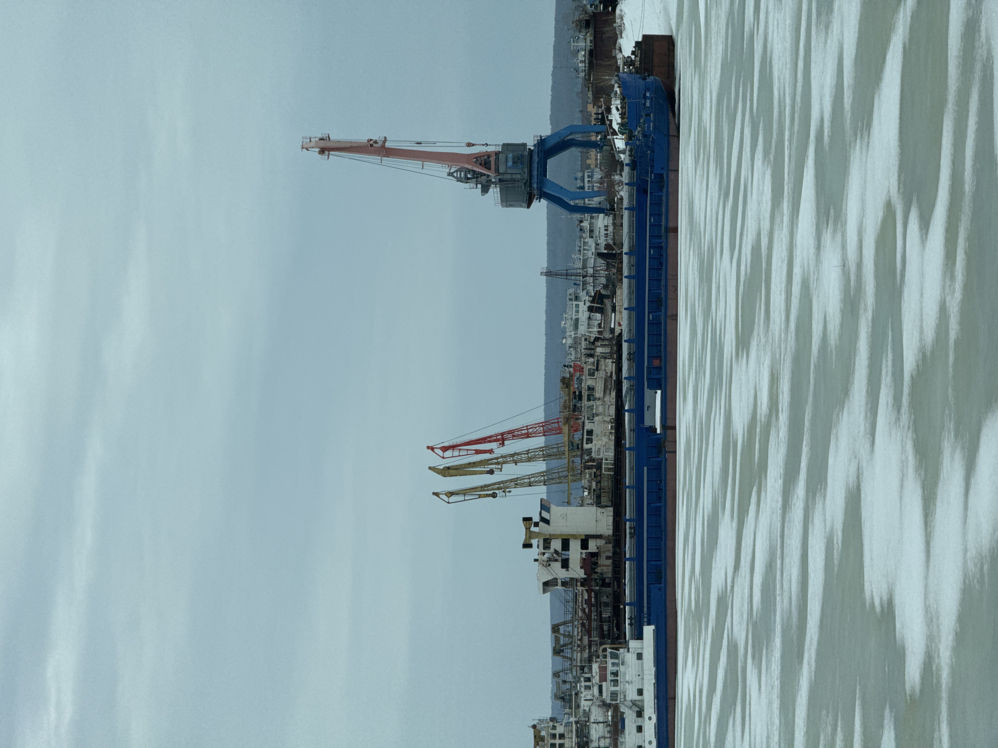 This image depicts a port area in Chaikovsky, Russia. Several cranes are visible along the dock, including a large blue and red crane prominent in the upper portion of the image, and smaller red and yellow cranes further down the dock. A blue-sided barge is moored alongside the dock. Other buildings and structures are present in the background, indicative of a developed urban area. The foreground features what appears to be a partially frozen body of water, with a pattern of light and dark areas on the surface. The sky is overcast.