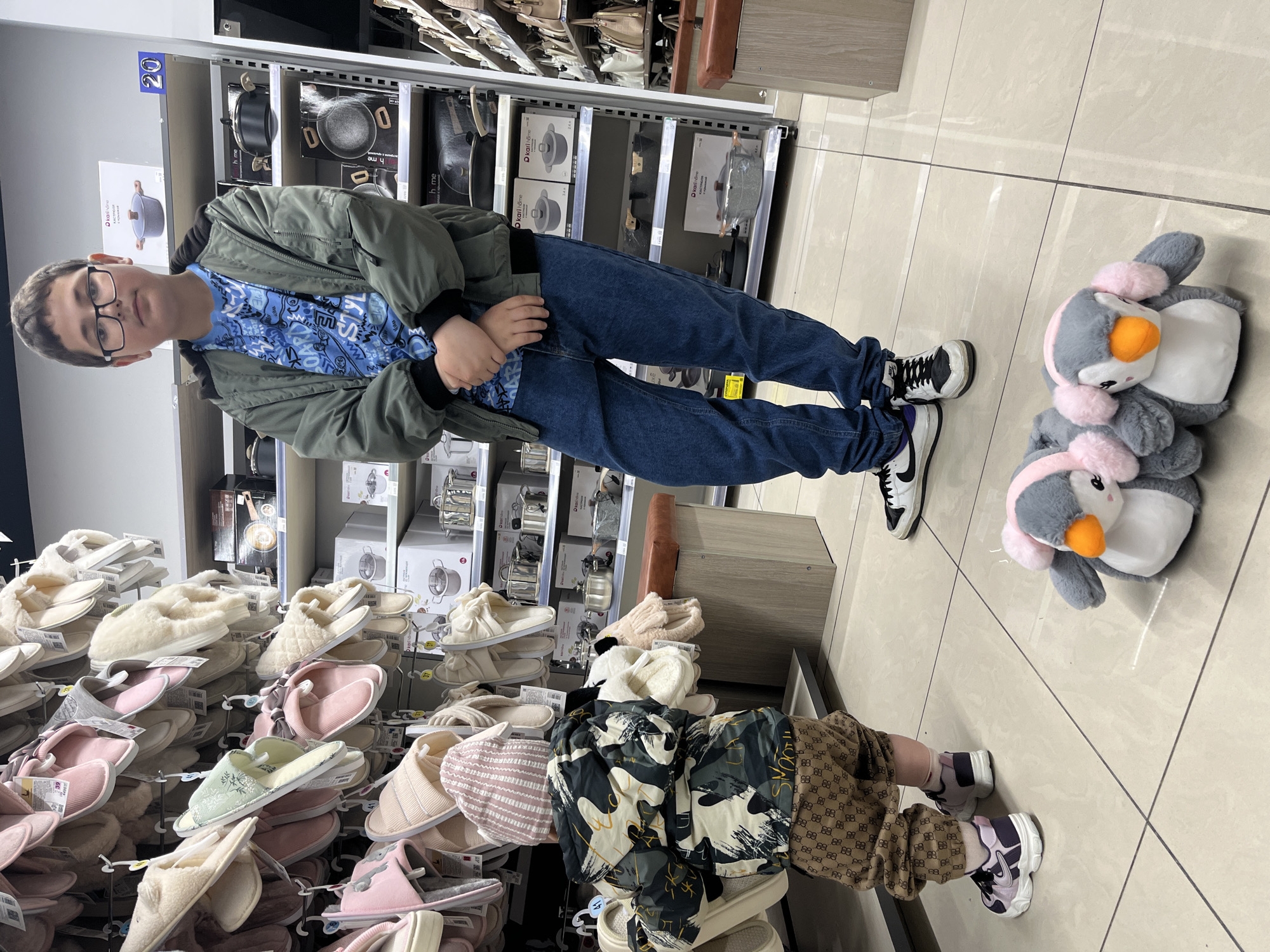 A photo shows a young boy in a green jacket and jeans standing in a store aisle in what is likely Sochi, Russia, though this is not confirmed by image content alone. He wears glasses and has his hands clasped in front of him. The aisle is stocked with household goods, including cookware and slippers, suggesting the store might be a department or home goods store. Additionally, there are plush penguin toys on the floor beside the boy. The composition and lighting suggest a casual snapshot taken indoors.
