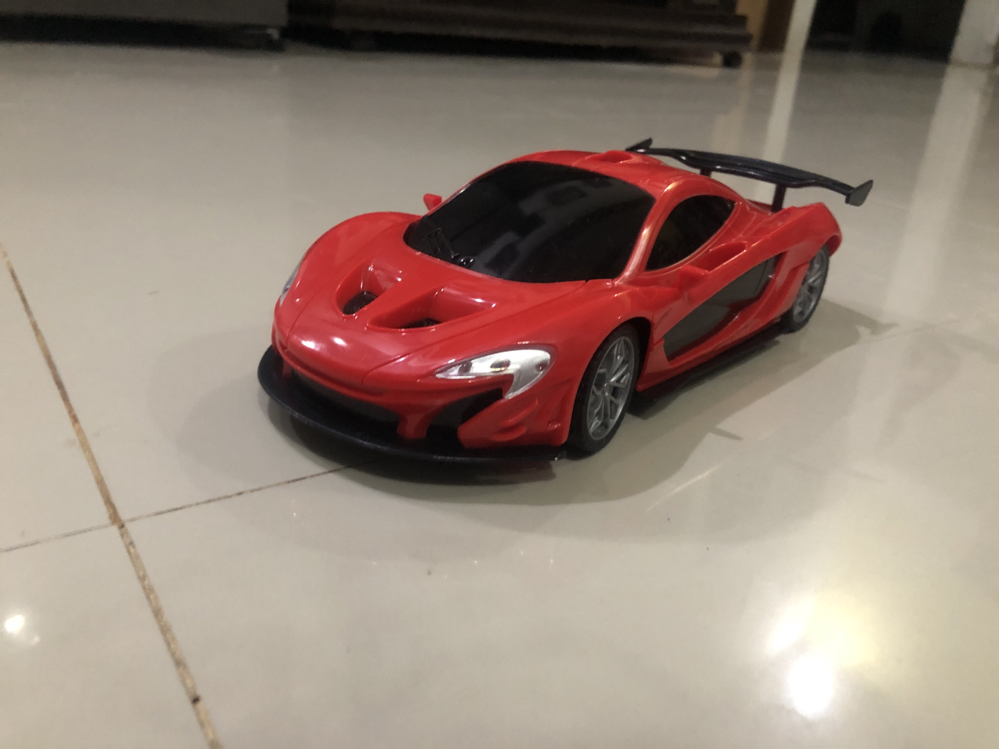 A red toy car sits on a light-colored tiled floor. The car is a model of a McLaren P1, with a black spoiler, black windshield, and silver wheels. The background is blurred, but shows a dark-colored object in the distance, suggesting the photo was taken indoors. The lighting is bright and reflects off the tiled floor. The location cannot be determined as Lhokseumawe, Indonesia from the image alone.