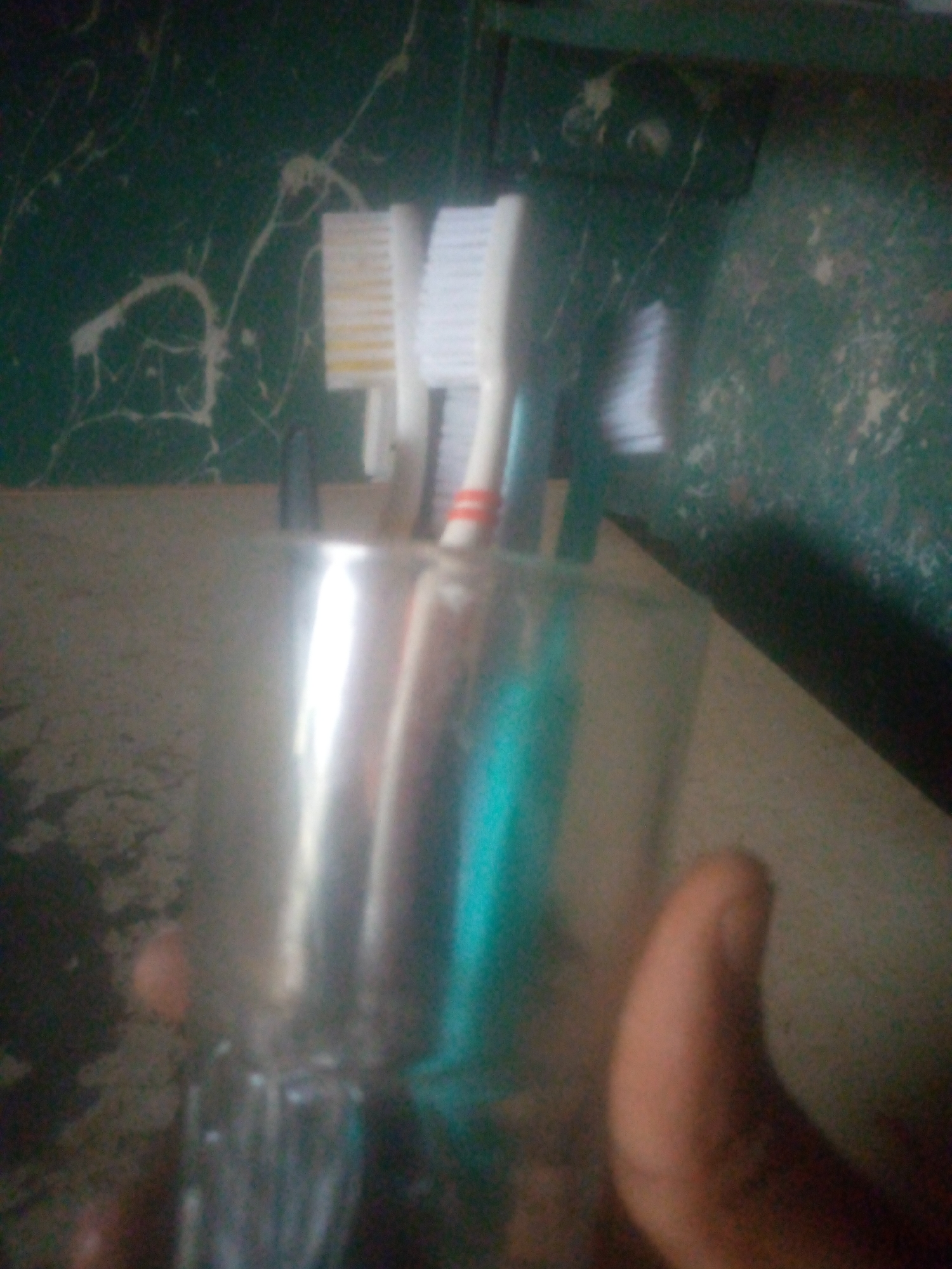The image shows a hand holding a clear glass with several toothbrushes inside. The toothbrushes have white bristles and colored handles. The background consists of a green surface with white patterns. The location is unknown.