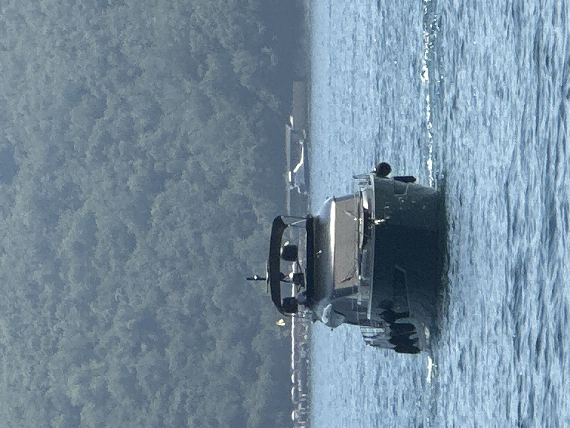 The image shows a motor yacht sailing on the water, with a lush, green hillside in the background. In the far distance, there are structures that appear to be part of a coastline, possibly with some buildings or piers. The water is a deep blue and appears slightly choppy, with small waves visible. The sky is overcast or hazy, lending a muted tone to the scene. The yacht is dark in color and has a covered cabin area. There are visible black fenders attached to the side of the yacht. Two figures are discernible within the cabin area of the yacht. In the background, on the water, there is another smaller vessel, possibly a speedboat or a personal watercraft, in the distance. The overall impression is a serene, outdoor scene, likely taken during the daytime. The location appears to be near Budva, Montenegro, given the characteristic mountainous coastline and the presence of yachts in the water.