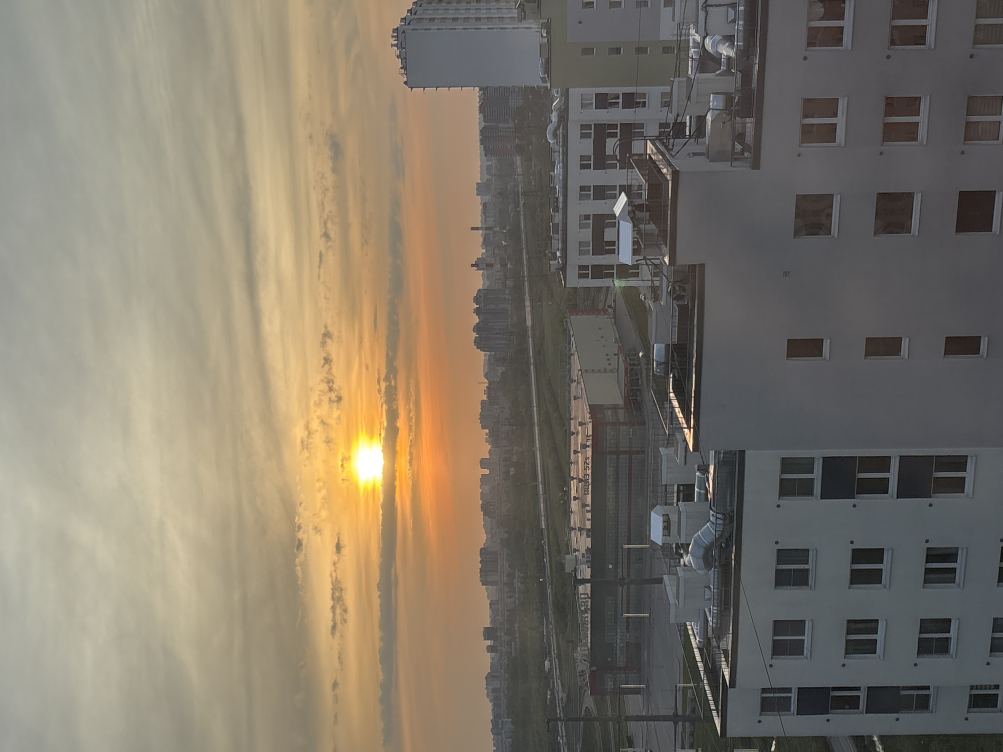 The image shows an urban landscape at sunset. On the left side of the frame is the sky, filled with clouds and a bright sun. In the middle, there is a cityscape including residential and commercial buildings. On the right is a building with many rectangular windows and some ducts. The image appears to have been taken from a high vantage point. The location is specified as Unknown, Russia.