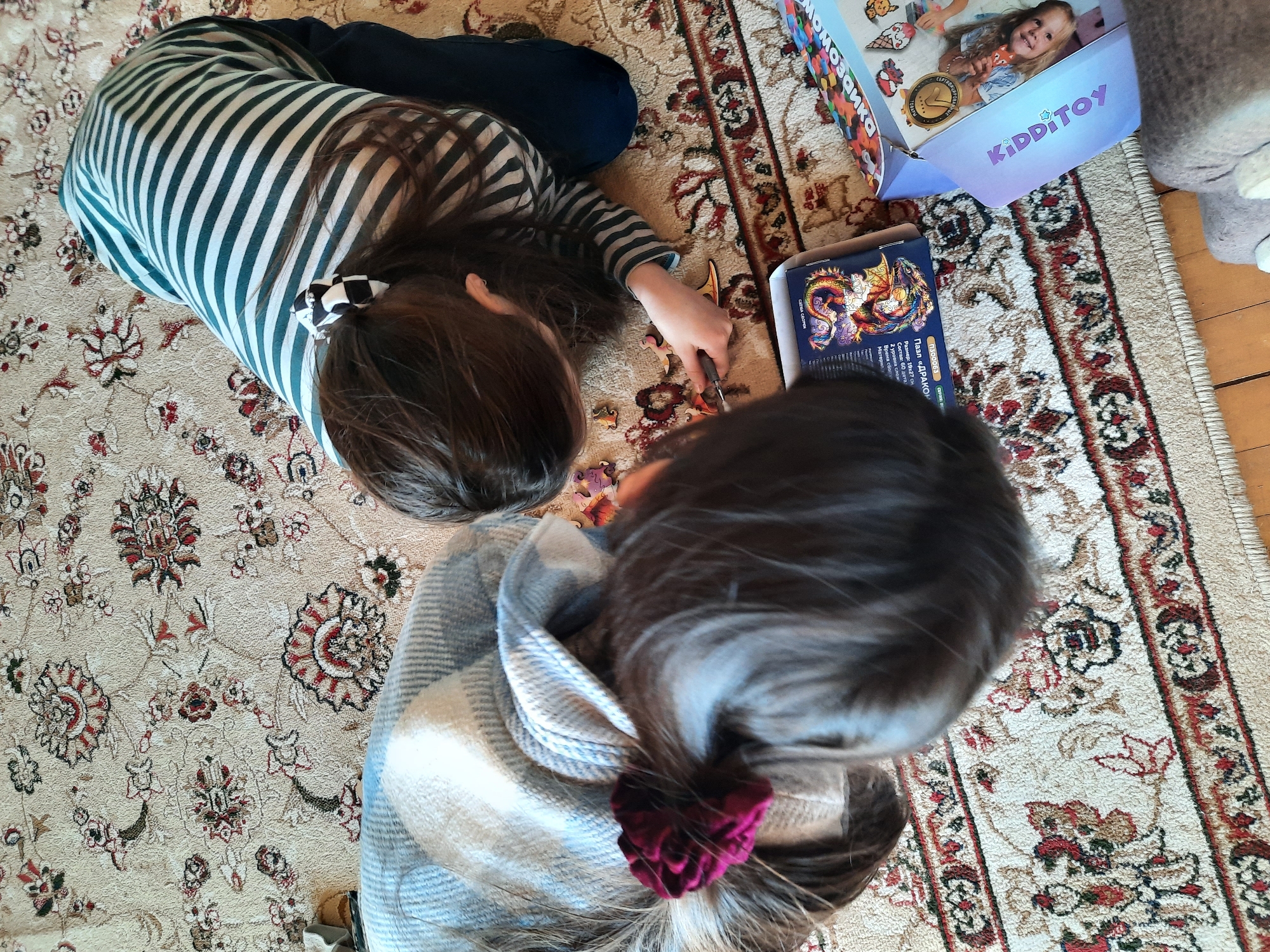 Two children are lying on a patterned rug indoors, engaged in putting together jigsaw puzzles. One child, wearing a striped shirt and black leggings, is working on a puzzle with a pair of tweezers. The other child is wearing a plaid shirt and has a burgundy hair scrunchie. Next to the children are two puzzle boxes, one labelled "KIDDITOY." A stuffed animal is partially visible in the upper right corner. The rug features a floral design with neutral, red, and black colors. A wooden floorboard is visible in the upper right corner. The location is not specified in the image.