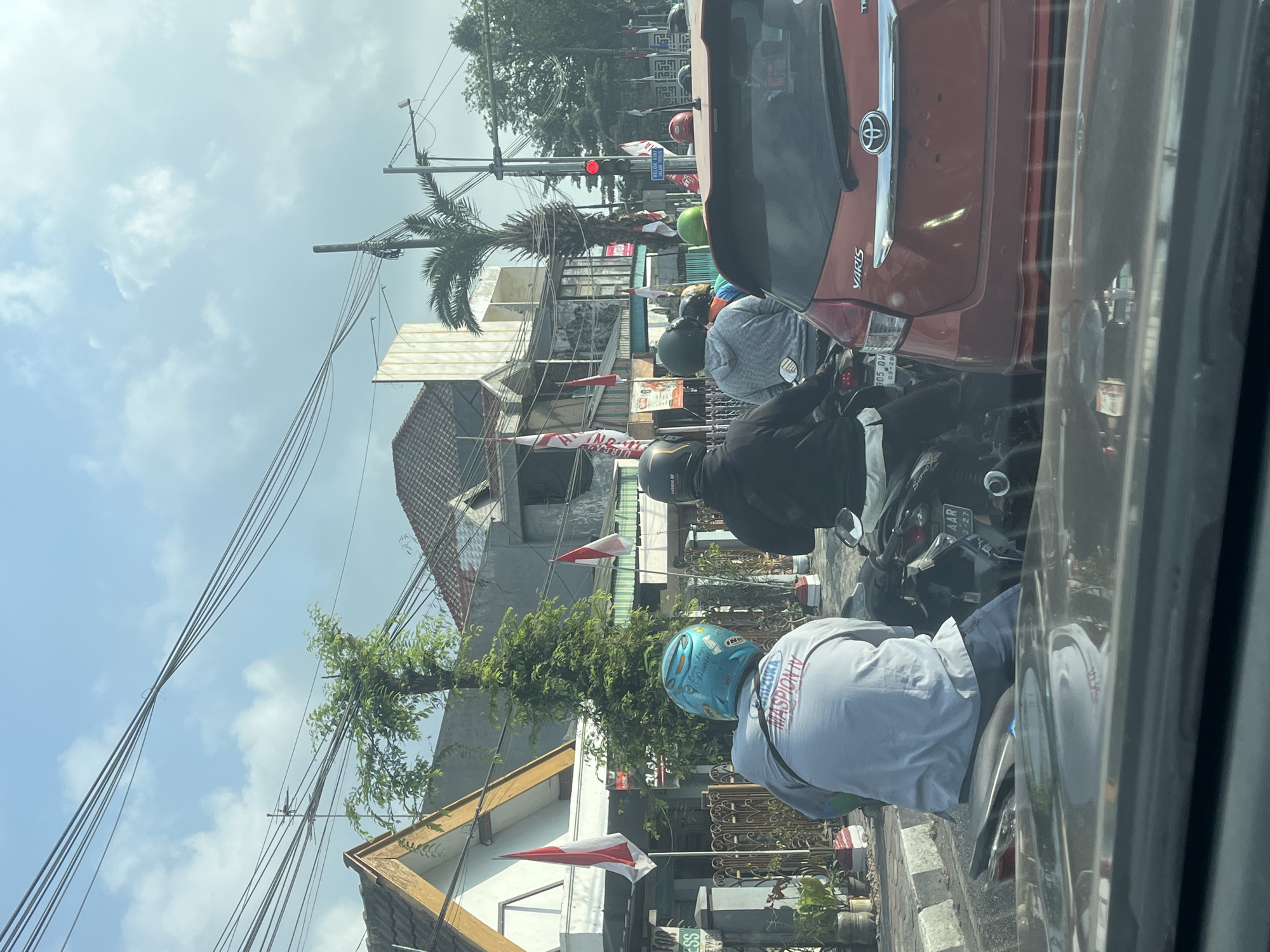 The image depicts a street scene in Gapurosukolilo, Indonesia, likely during daytime given the bright sky and shadows. The foreground shows the dashboard and windshield of a car. Behind it, traffic is visible, primarily consisting of motorcycles. A person wearing a light blue helmet and a white long-sleeved shirt is seated on a motorcycle, facing away from the camera. To their right, another person in a black jacket and dark helmet is also on a motorcycle. Further ahead, a red Toyota Yaris is stopped.
The background reveals buildings with red-tiled roofs and some with white facades. Indonesian flags, red and white, are prominently displayed on poles outside some of the buildings. A traffic light, showing red, is visible on the right side of the street. Several power lines crisscross the sky. Trees, including a palm tree, are present along the street. The overall impression is of a typical urban street with moderate traffic and clear weather.