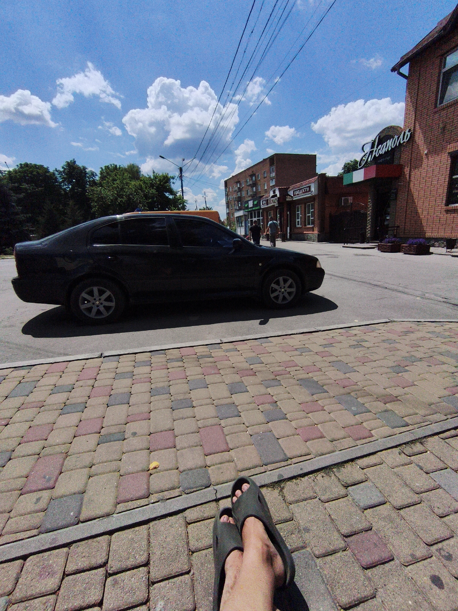 A low-angle, wide shot captures a street scene under a bright, cloudy sky in Velyka Bahachka, Ukraine. In the foreground, a person's bare feet in dark flip-flops rest on a patterned paved walkway composed of interlocking bricks in shades of red, gray, and beige. The walkway is bordered by a gray concrete strip. Beyond the walkway, a black car is parked on the right side of the asphalt road. The car, a sedan, casts a shadow on the road. To the right of the car, a two-story brick building with arched windows and a sign that reads "L'canolo" in ornate script is visible. A stripe of the Italian flag colors, green, white, and red, is displayed below the sign. Further down the street, a multi-story brick apartment building with numerous windows stands. A few trees line the left side of the street, and utility poles with wires crisscross the sky. Two figures, likely pedestrians, are walking away from the camera on the sidewalk in front of the apartment building. The overall impression is a sunny day in a semi-urban environment.