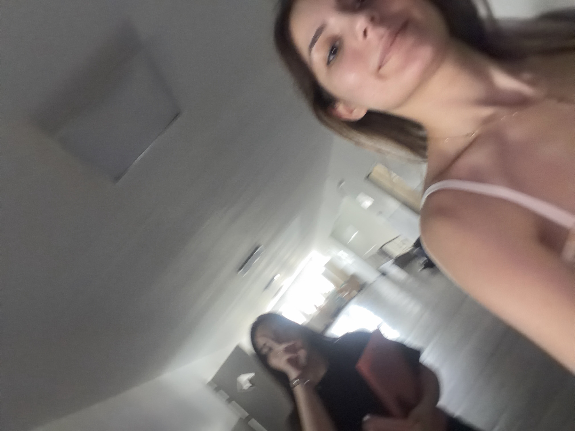 An indoor, low-angle shot captures two young women in what appears to be a hallway or large room in Burgas, Bulgaria. In the foreground, positioned on the right and slightly out of focus, is a woman with light brown hair, wearing a light-colored, possibly white or light pink, spaghetti strap top and a delicate necklace. She has a subtle smile on her face, with her gaze directed slightly upwards.
Further back and to the left, significantly blurred, is another woman with dark hair. She is wearing a dark top and is holding a reddish-orange object, possibly a book or tablet. One of her hands is near her face, and she appears to be looking downwards or away from the camera.
The setting features light-colored walls and ceiling, with a square light fixture visible on the ceiling in the upper left. The floor is a consistent dark tone. Bright light streams in from the background, creating some glare and making parts of the background overexposed, suggesting a daytime setting or a brightly lit interior. The overall impression is a casual, possibly selfie-style, photograph of two individuals in an informal indoor environment. No text is visible in the image.