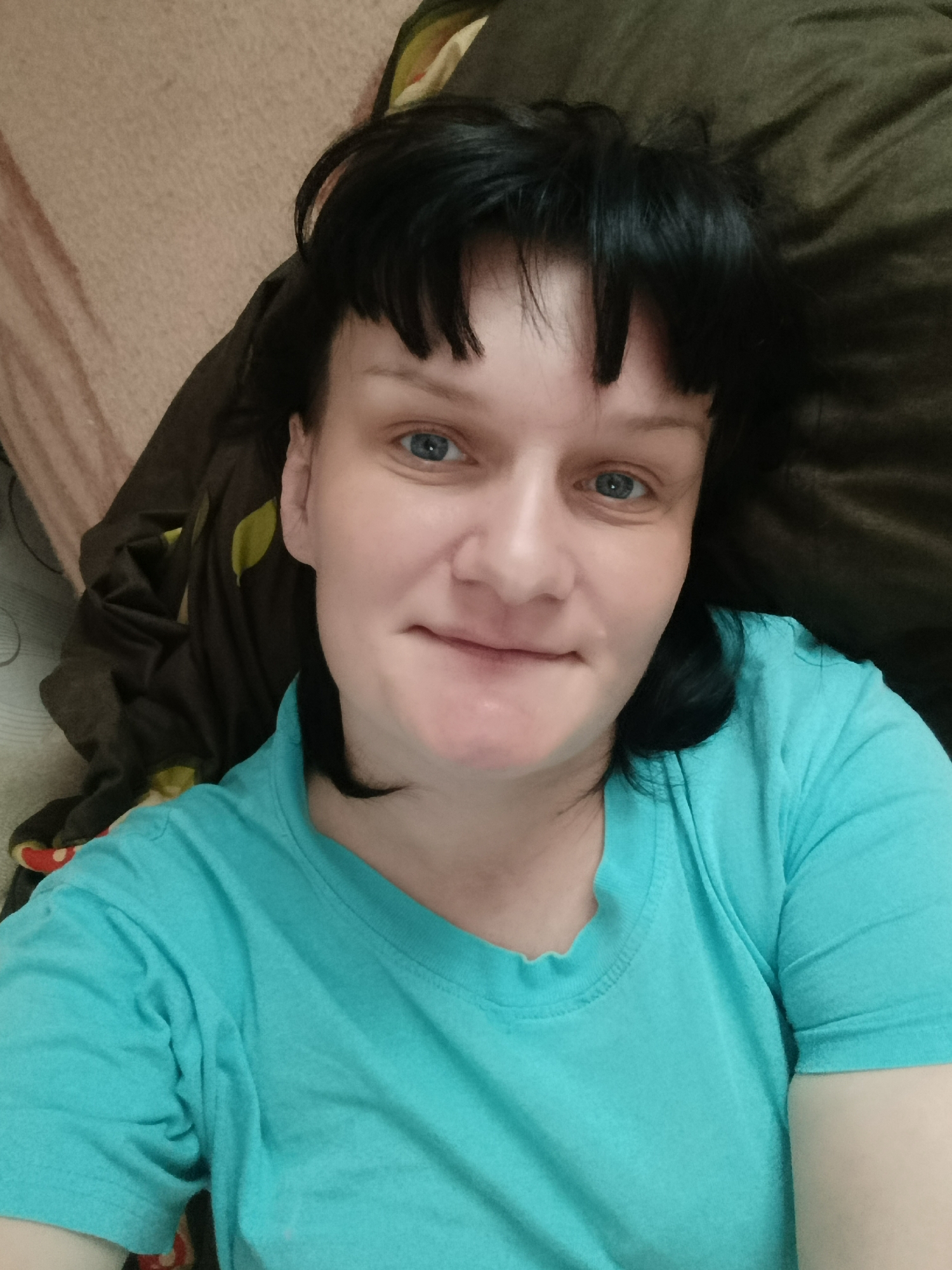 A woman with fair skin, blue eyes, and dark hair with bangs is lying down. She is wearing a teal t-shirt. The background includes brown and tan fabrics, potentially bedding. The location, Фоки, Russia, is not evident within the visual content of the image.