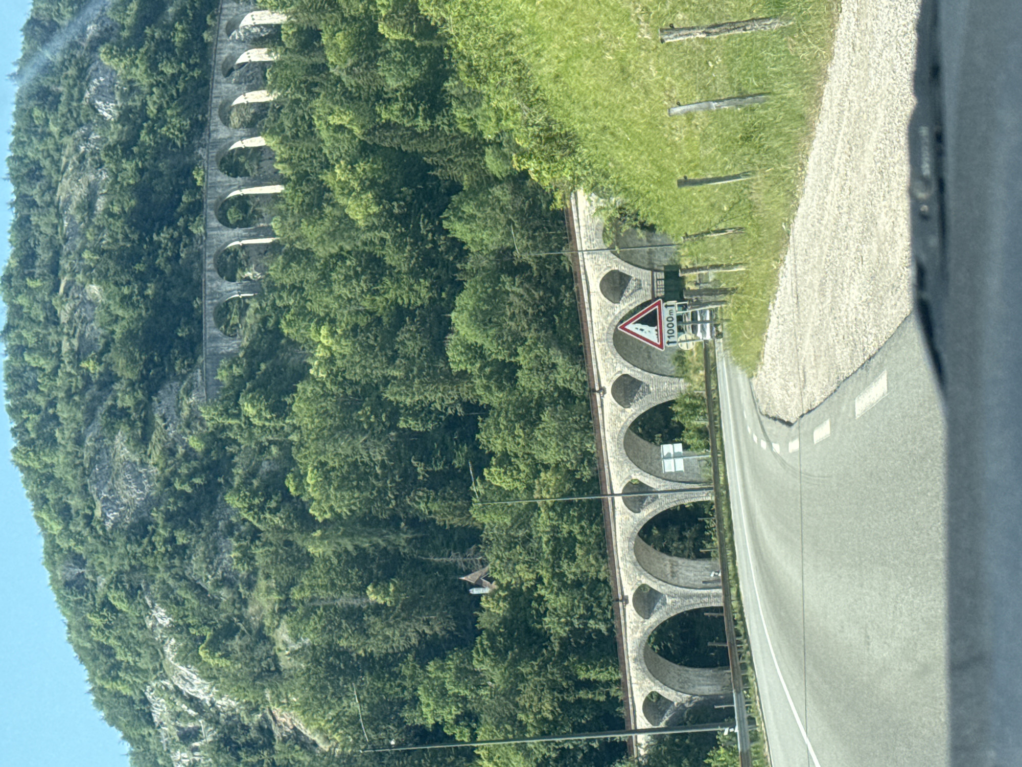 This is a photograph of the Oigney Viaduct in the city of Hauts de Bienne, France. The viaduct is made of stone arches and spans across a green, forested mountainside. In the foreground, a road curves to the right, with a grassy embankment on the right side of the road. There is a traffic sign with a mountain and "1000m" on it. In the background, the mountainside is covered in trees. The sky is clear and blue.