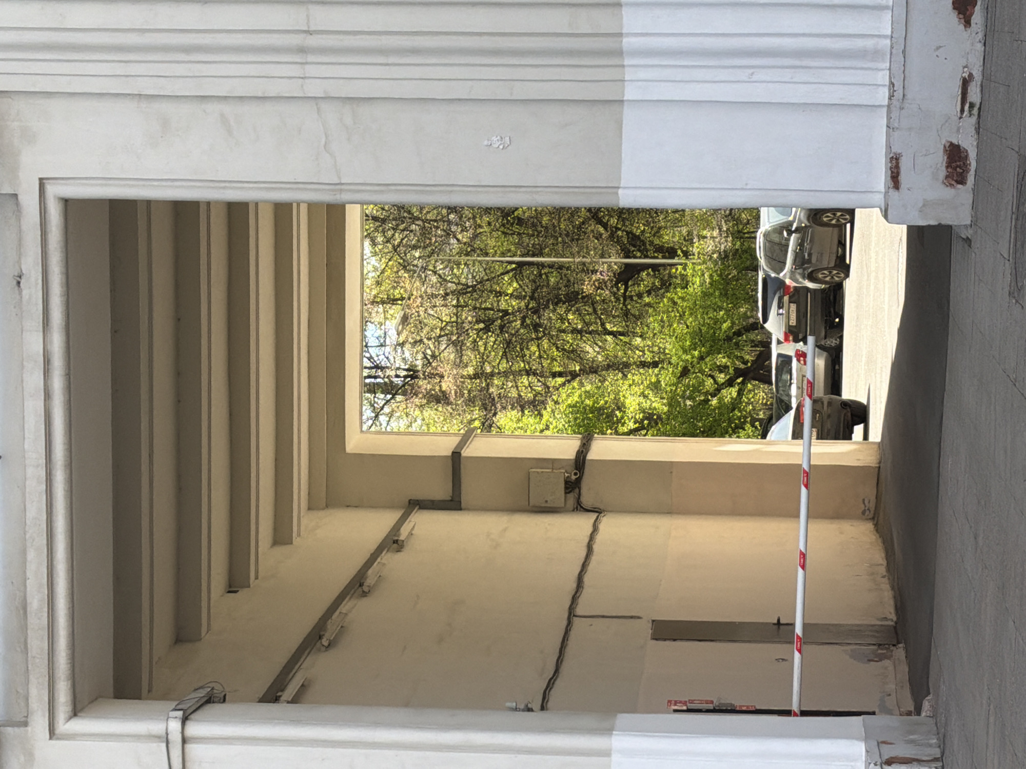 The primary subject is an architectural passageway or archway, possibly an entrance to a building or courtyard, seen from within. The passage is made of light-colored, possibly concrete or stone, material. Through the opening, a street scene is visible with parked cars and lush green trees.
The most prominent activity suggested is renovation or construction. The upper part of the archway (or molding) shows a clear division between an older, off-white, and somewhat weathered section on the left and a freshly painted, bright white section on the right. Inside the passage, electrical wires run along the upper wall/ceiling, connected to a small white junction box. A white and red striped pole, resembling a barrier or construction marker, stands upright near the far side of the passage opening.
The scene is set outdoors during bright daytime, under what appears to be a clear, sunny sky, indicated by the strong light and shadows. The trees visible through the archway are full of green leaves, suggesting spring or summer. The cars parked outside appear to have Russian license plates (e.g., partial plate "X377KO" on a dark sedan). The interior of the passage has vertical, rib-like architectural elements along its left wall. The floor of the passage shows some unevenness and possibly loose construction materials or debris. The overall impression is an urban environment undergoing maintenance or aesthetic renewal in Moscow, Russia.
The white and red striped pole has multiple red rectangular stickers with white text, some of which appear to read "T.D.A." and others are illegible due to resolution or angle.