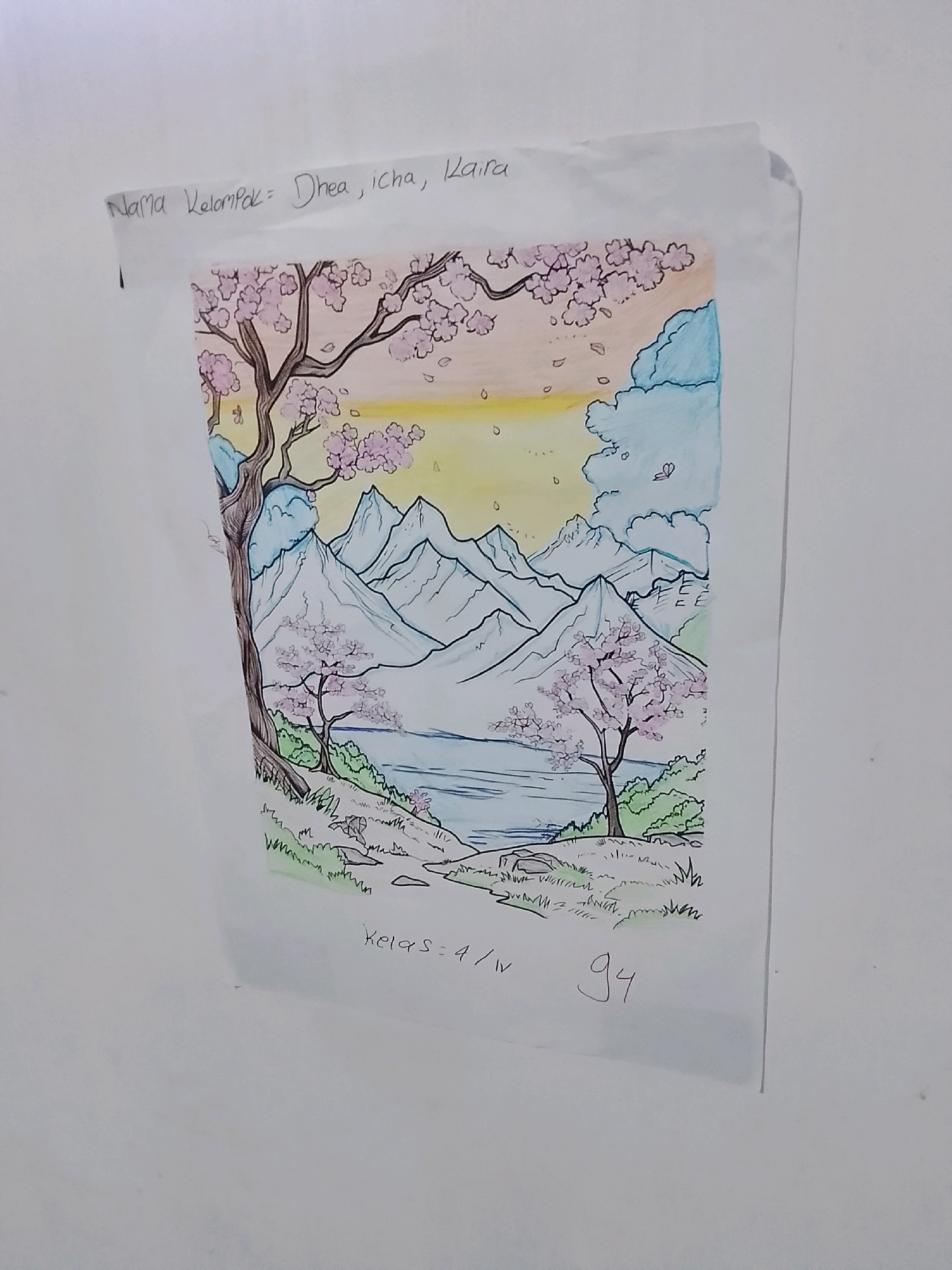 A hand-drawn picture of a landscape is taped to a white wall. The drawing depicts a serene scene with snow-capped mountains in the background, a calm lake in the middle ground, and cherry blossom trees in the foreground. The sky is a gradient of yellow and light orange, with some blue clouds. Text is written at the top of the drawing that reads "Nama Kelompok = Dhea, Icha, Kaira". Below the drawing, text reads "Kelas = 4/IV" and a number "94". The image is taken from a slight angle, suggesting it is a photograph of the artwork. The location is specified as Ujungrusi, Indonesia.