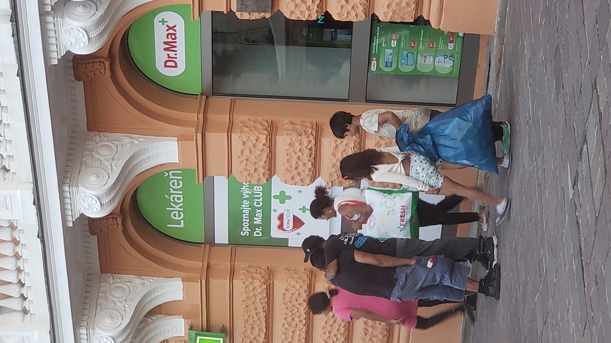In Prešov, Slovakia, on a cobblestone street, a group of people stand outside a Dr. Max pharmacy. A woman in a pink dress walks away from the camera on the left. Next to her, two men embrace, with one wearing a black baseball cap. A woman in a white top and skirt stands holding a large white grocery bag with the word "FRESH" printed on it. Behind her, a woman with dark hair wears a white top and patterned shorts. To her right, a young boy in a light-colored t-shirt stands. In front of the boy, a person is obscured by a large blue plastic bag. The pharmacy's green signage displays "Lékáreň" and "Dr. Max+". Another section of the pharmacy's window shows product information, with "Dr. Max" visible. The building's facade is textured and painted orange, with decorative white moldings around the arched doorways.