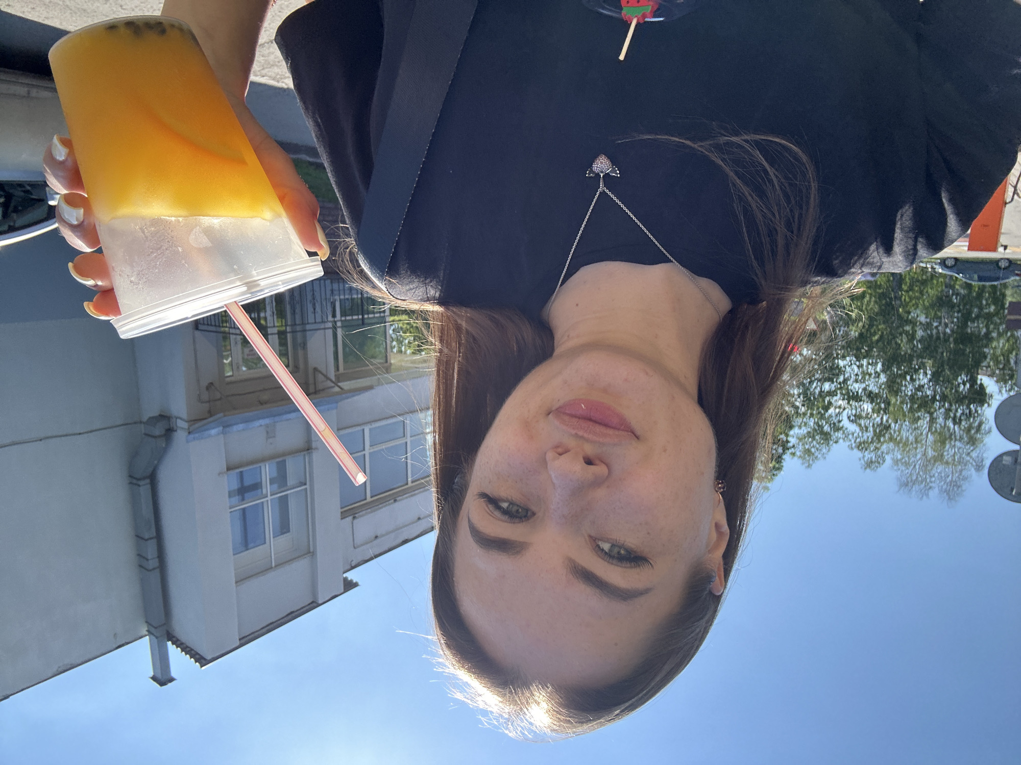 A young woman with brown hair and freckles is holding a clear plastic cup with a yellow beverage and a pink straw. She is wearing a black t-shirt and a silver necklace with a heart-shaped pendant. The woman is outdoors, with a building and trees in the background. The sky is clear and blue.