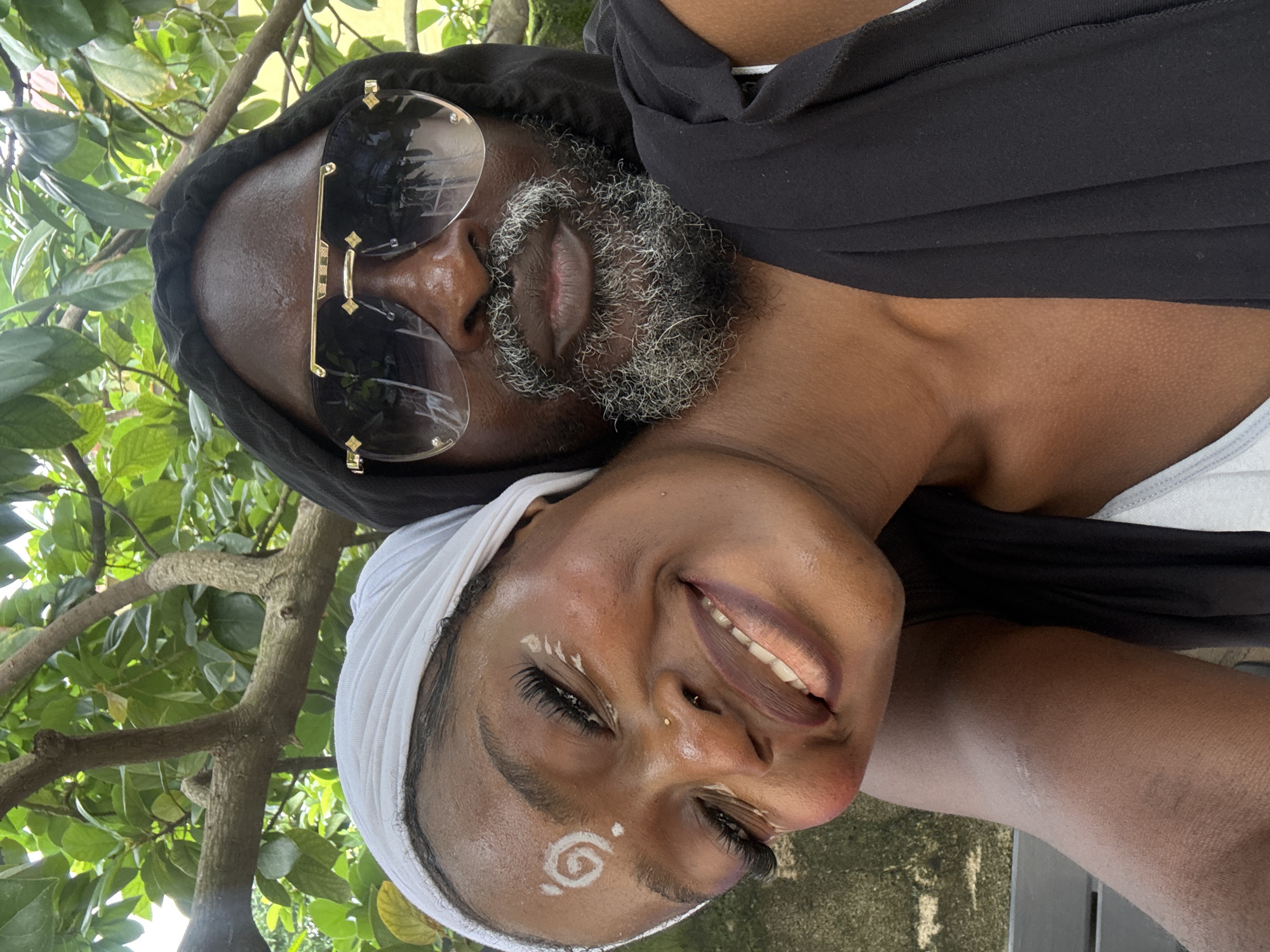 In Ado, Nigeria, a close-up, eye-level selfie shows two individuals posing outdoors under leafy tree branches. The person in the foreground, on the left, is a young Black woman wearing a white headscarf and adorned with white face paint, including a spiral design on her forehead and white markings around her eyes and cheeks. She is smiling broadly, looking towards the camera, with her eyes closed and her head tilted. The second individual, a Black man, is positioned behind and to the right of the woman, wearing a black hooded garment and large, dark sunglasses with gold accents. He has a salt-and-pepper beard and mustache and is also looking towards the camera. The background consists of blurred green foliage from the trees, with hints of a concrete wall and a wooden bench visible.