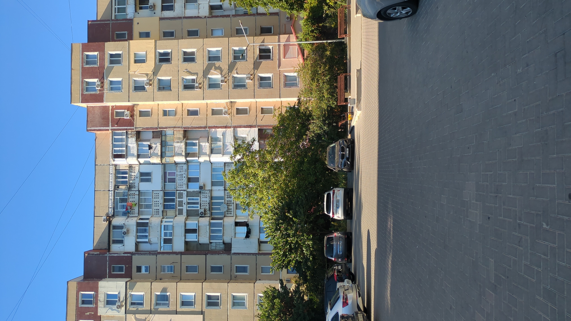 The image depicts a street scene in Chișinău, Moldova, featuring a large, multi-story apartment building under a clear blue sky. The building has a facade that is a mix of beige, light brown, and maroon sections, with numerous windows and balconies. Many balconies are enclosed, and some have air conditioning units mounted on them. The ground level of the building appears to be residential or commercial.
In the foreground, a paved area, likely a parking lot or courtyard, is visible, made of interlocking rectangular grey pavers. Several cars are parked on this paved surface. To the left, a white SUV is parked. Next to it, a darker SUV is parked, followed by a grey car. In the center of the frame, a white SUV and a dark grey sedan are parked. A portion of a silver car is visible on the far right edge of the image.
Between the parking area and the building, there is a strip of greenery with several mature trees and bushes. Two wooden park benches are placed in this green area, facing the parking lot. A street lamp stands near the right side of the image, adjacent to the building. The overall impression is of a residential urban environment on a sunny day.