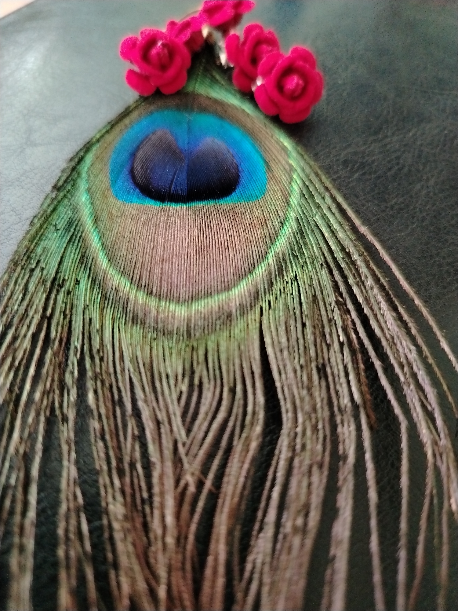 The image displays a close-up of a peacock feather on a black, textured surface, likely leather. Three small, fuzzy, bright pink decorative flowers are attached to the top of the feather. The peacock feather exhibits its characteristic eye-like marking with vibrant blue and green hues. The rest of the feather consists of fine, elongated barbs in shades of brown and green. The overall lighting is soft, highlighting the textures and colors of the objects. There is no visible information in the image to indicate a specific location in Krasnodar, Russia.