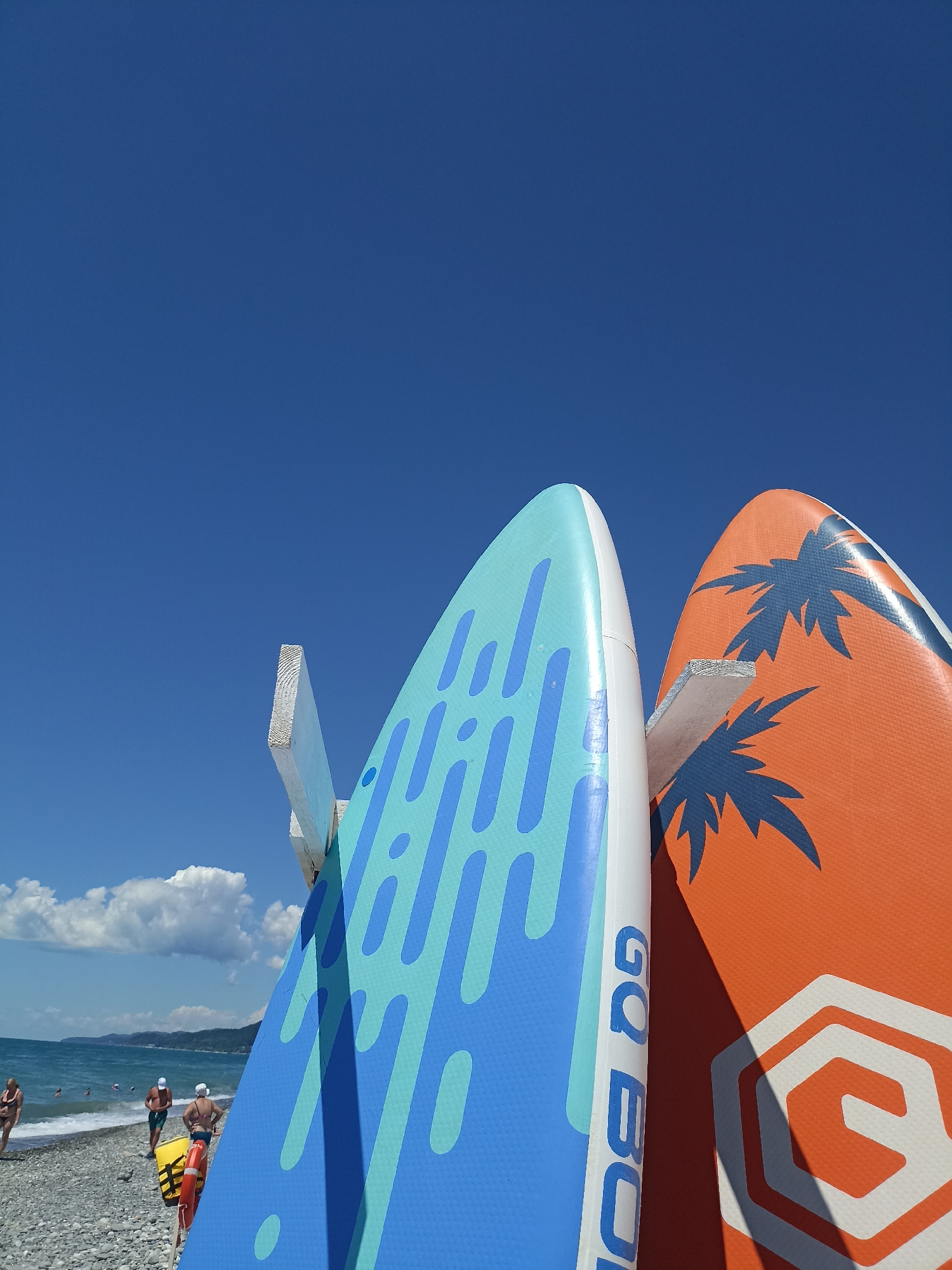 The image depicts a sunny day at a pebble beach in Sochi, Russia. Two stand-up paddleboards are prominently displayed in the foreground, leaning against a white wooden support. The paddleboard on the left is predominantly light blue with a pattern of darker blue rounded shapes and lines. The paddleboard on the right is bright orange with a stylized navy blue palm tree design and a white hexagonal logo. The words "GO BO..." are visible vertically on the side of the left paddleboard.
In the background, the Black Sea stretches out under a clear, vibrant blue sky with scattered white clouds. The shoreline is lined with people, some of whom appear to be preparing for or returning from water activities. A few individuals are visible in the water. The beach is covered in gray pebbles. The overall impression is one of a recreational summer day at the seaside.