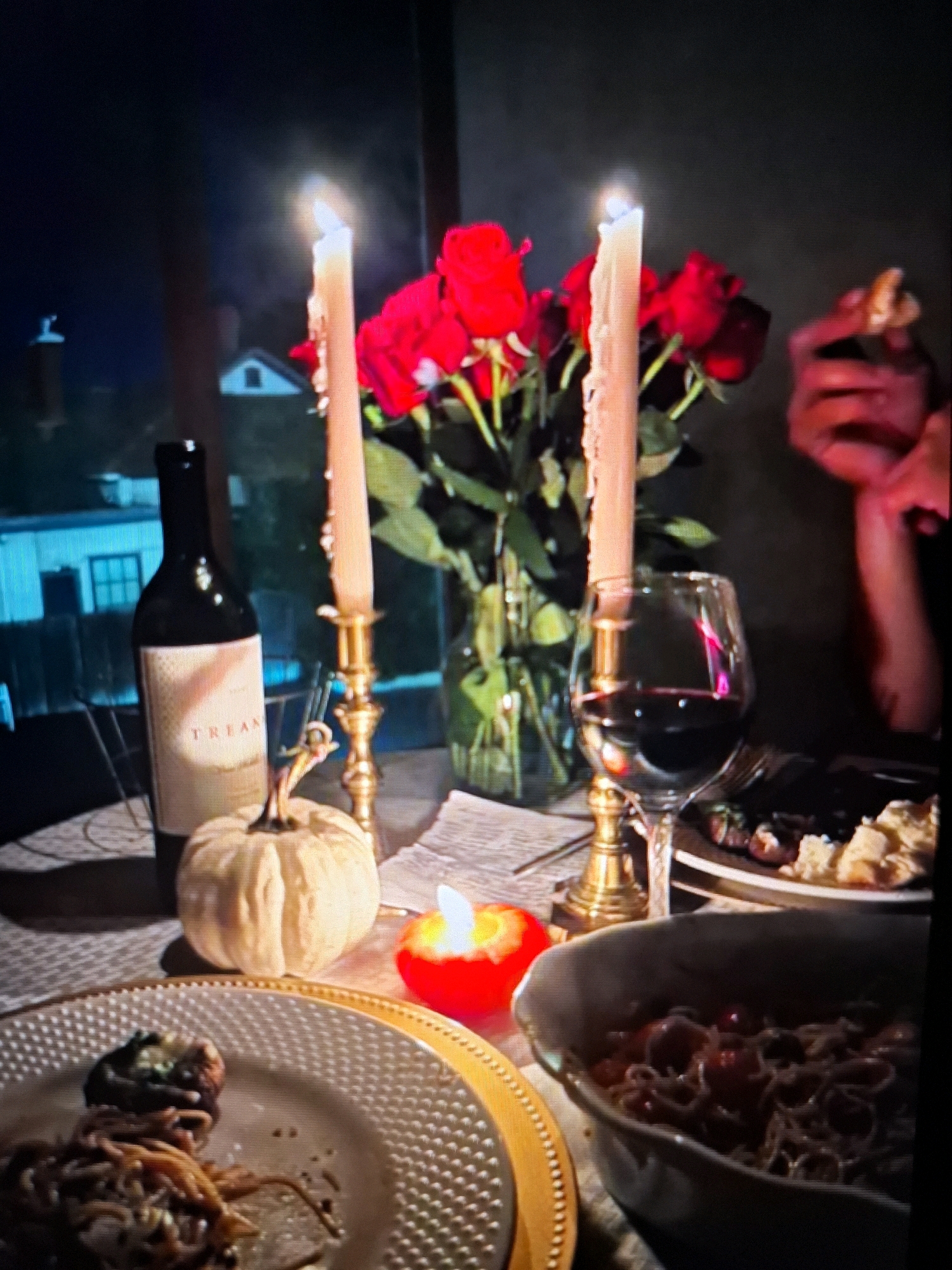 The image depicts a dimly lit, intimate setting, likely an indoor dining area, with a romantic ambiance. In the foreground, a plate of spaghetti is visible, partially obscured by a serving dish containing more spaghetti and what appear to be cherry tomatoes. To the left of the plate, a decorative white pumpkin sits on a textured placemat. A bottle of red wine, labeled "TREAM," stands upright next to a brass candleholder with a lit, tapered candle. Two tall, lit candles are prominent, their flames casting a warm glow on a bouquet of vibrant red roses in a glass vase situated behind them. A wine glass filled with red wine is positioned to the right of the candles, reflecting the candlelight. In the background, the faint outlines of buildings and trees suggest an outdoor environment visible through a window. A person's hand, holding a piece of food, is visible in the upper right quadrant of the image, interacting with the scene. The overall impression is of a prepared meal for a special occasion. The image does not provide any specific visual cues or landmarks that would allow for its identification within the city of Yamunanagar, India.
