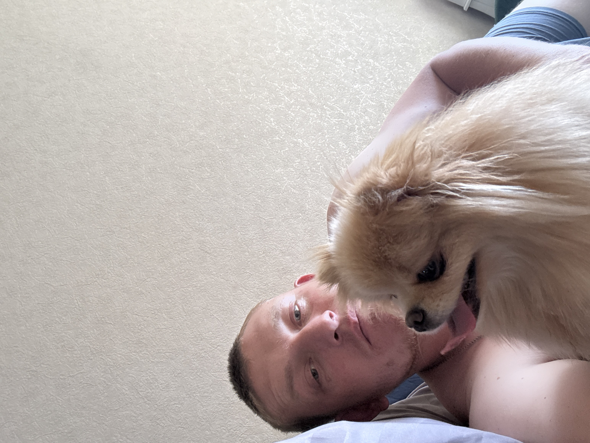 A man and a dog are pictured from the chest up in a close-up shot. The man is on the left side of the image, looking directly at the camera with his head slightly tilted. He has short brown hair and is shirtless, wearing a necklace. A small, fluffy dog with light brown fur is held in his arms, facing away from the camera and licking the man's face. The background is a textured, off-white wall. The lighting is soft and natural, suggesting an indoor setting.