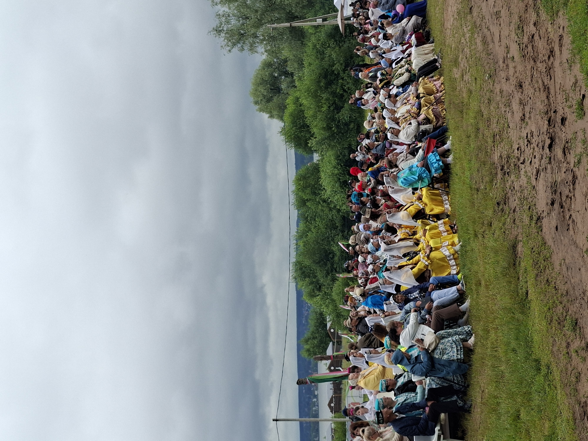 The image shows a crowd of people, mostly women, seated on benches outdoors, attending an event. Many of the women are wearing traditional attire, including headscarves and colorful dresses. The setting appears to be a grassy field with trees and a body of water in the background. The sky is overcast, suggesting a cloudy day. The people are seated facing away from the camera, indicating they are watching something in front of them. The ground in the foreground is a mix of grass and dirt. The image does not contain enough information to determine the specific location within Votkinsk, Russia, other than it is an outdoor public gathering.