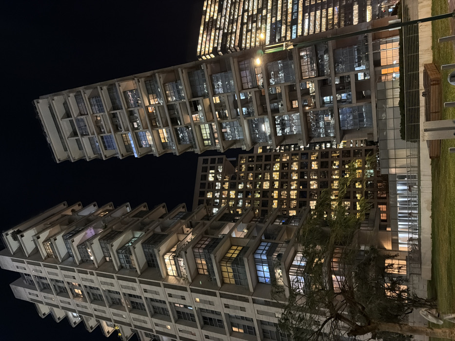 Tall buildings with lit windows dominate the night scene. The architecture features projecting balconies and staggered designs. A grassy area with a few benches is visible on the right. A sparse tree stands in front of the buildings, partially obscuring the lower floors. The sky is dark, contrasting with the illuminated windows.