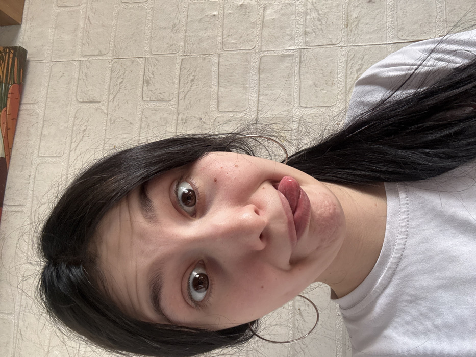 The image displays a young female, situated indoors within the city of Фоки, Russia. She is centrally positioned, looking directly into the camera with wide-open eyes and raised eyebrows. Her dark, long hair is parted and pulled back, with a portion resting over her left shoulder. She wears a plain white t-shirt and large, circular, silver-colored hoop earrings. Her mouth is contorted in a playful expression, with her tongue extended downwards and to the viewer's left. The background consists of a light-colored wall with a textured, brick-like pattern. A framed object depicting stylized carrots and green leaves is partially visible in the upper right corner of the frame.
