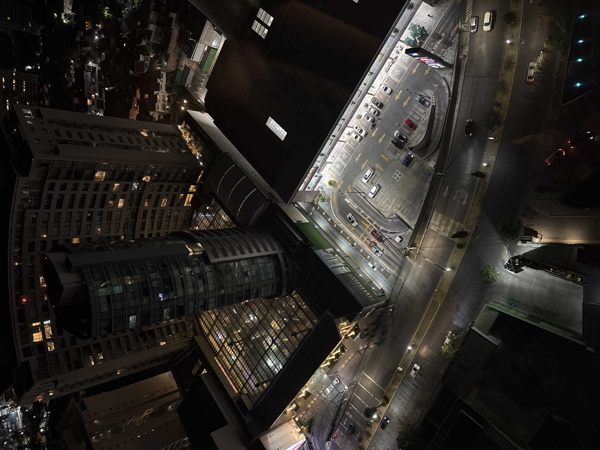 The image presents an aerial view at night, capturing a blend of urban infrastructure and residential areas in Jesús del Monte, Mexico. The foreground is dominated by modern architecture, specifically a high-rise building with a distinct curved glass façade. Adjacent to this building is another structure featuring a grid-like glass exterior, illuminated from within, creating a patterned light effect.
Beyond the building complex, the view extends to a well-lit parking area with clearly marked parking spaces and directional arrows. Several vehicles are parked in this area. The adjacent road is also illuminated, with visible traffic, including cars moving along the road. Streetlights line the road and parking area, contributing to the overall brightness of the scene.
In the background, a network of dimly lit residential areas is visible. Distant lights from homes and buildings are scattered across the landscape, providing a contrast to the brighter, more structured lights of the commercial and parking areas. The overall scene suggests a mix of residential and commercial activity.