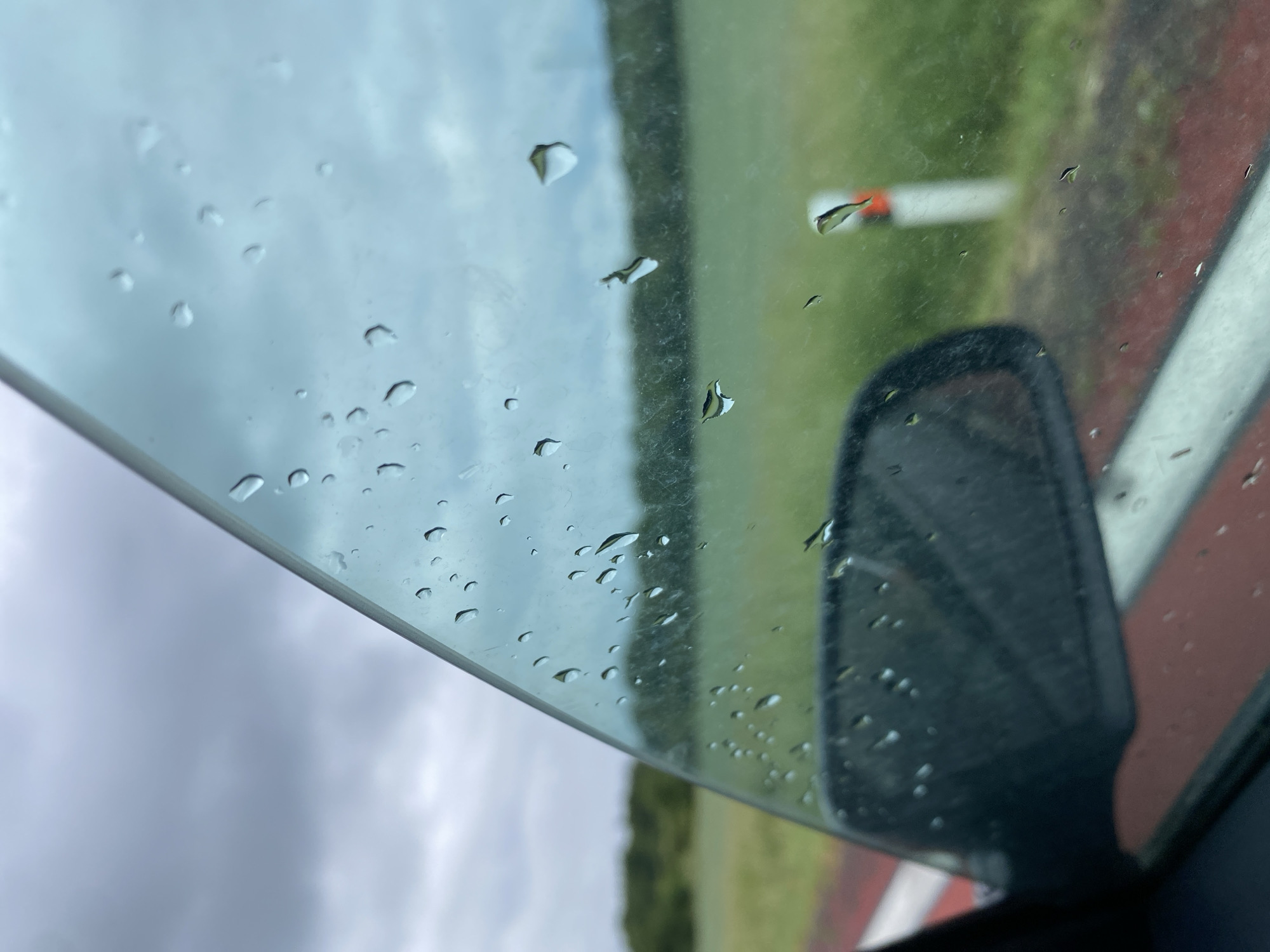 The image, taken from inside a vehicle, offers a view through a rain-streaked car window. The foreground shows the edge of the car's side mirror and the red and white stripe of the car door frame. The window itself is covered in numerous water droplets, distorting the view of the outside. Beyond the window, a blurred landscape unfolds, characterized by a green field or pasture, a line of trees in the mid-ground, and a cloudy, overcast sky. A white post with an orange band is visible in the distance, suggesting a roadside or open area. The overall impression is of a cool, possibly damp day, with limited visibility due to the rain. The location is stated to be Mirošovice, Czechia. There are no people or animals visible in the image.