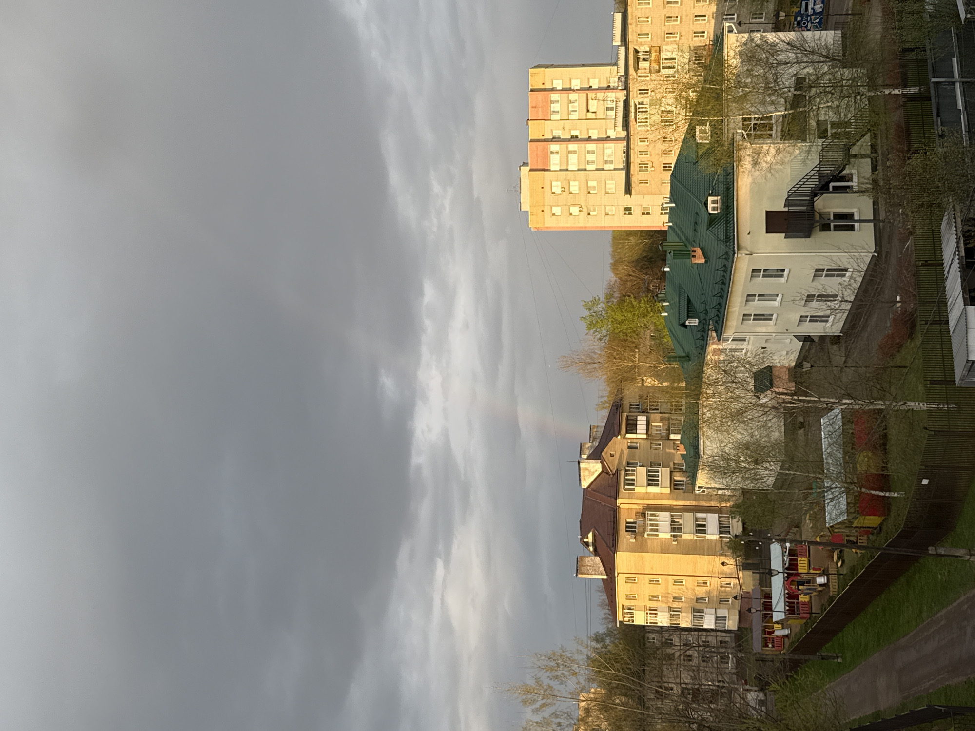 The image captures an urban landscape in Rusanovo, Russia, featuring apartment buildings under a cloudy sky with a faint rainbow arc visible. The architecture includes light-colored, multi-story residential buildings with numerous windows. Several structures have green roofs. The ground level reveals trees with budding leaves, a playground with colorful equipment, a fence, and a paved walkway. The overall light suggests it's either early morning or late afternoon.