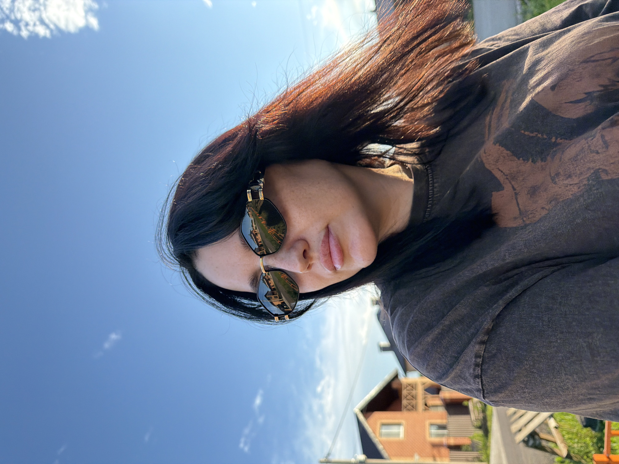 A woman with dark hair and sunglasses is outdoors in what appears to be a suburban area, possibly the village of Shmidotovo, Russia. She is wearing a dark, graphic t-shirt. The background features a clear blue sky with a few wispy clouds, and houses with red brick exteriors. The sun is shining, casting a warm light, and her hair is being blown by the wind. The reflection in her sunglasses shows more of the houses and the sky.