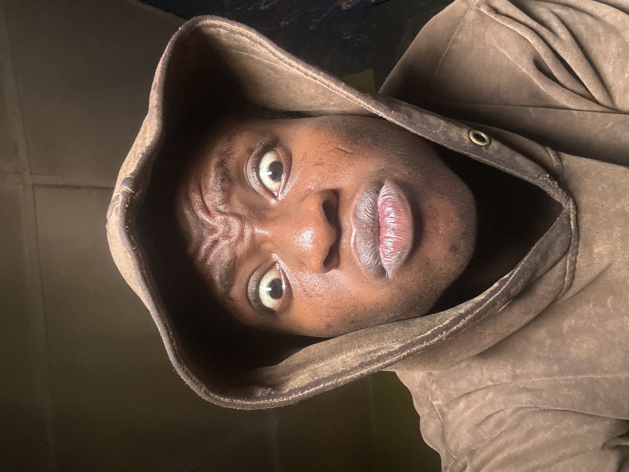 A medium shot captures a Black male with a deeply furrowed brow, appearing apprehensive or concerned. He is enclosed within a brown hooded sweatshirt, the hood framing his face. The background is dimly lit. The location is Unknown, Unknown.