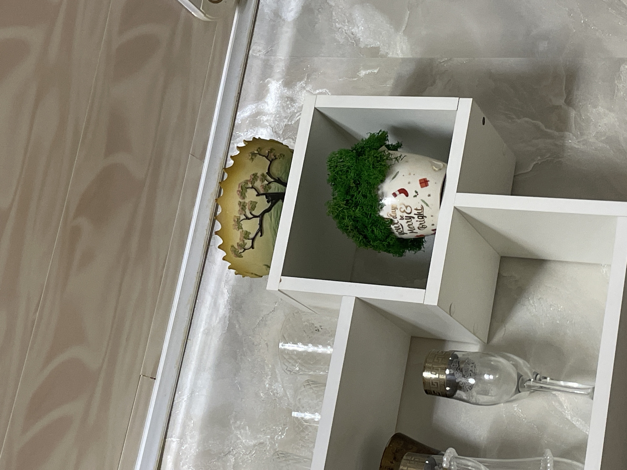 The image shows a close-up of a shelf unit with decorative items. On the top shelf, there is a yellow, scalloped dish with a painted image of a tree. Below this, on a higher shelf of the unit, are two clear glass tumblers with a diamond pattern. On the shelf below, in a white cubic compartment, is a white ceramic pot filled with a dense clump of green moss. The pot has festive Christmas-themed decorations, including the words "your day be Merry & Bright," along with images of stockings and gift boxes. The background wall has a textured, marbled appearance in shades of gray and white. The shelf unit itself is white. The overall scene is indoors, suggesting a home or retail environment, possibly a kitchen or living area. The image does not contain any information to indicate it is located in Laishevo, Russia.