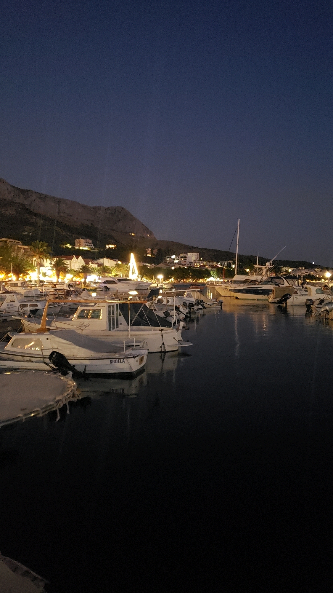 In the city of Općina Tučepi, Croatia, a nighttime scene unfolds at a harbor filled with docked boats. The sky is a deep twilight blue, with the faint glow of lights from a town nestled against a mountainous backdrop. Several boats, varying in size and type, are moored in the calm water, their forms illuminated by a combination of their own lights and the ambient town lights. One boat in the foreground has the name "Sardela" visible on its hull. The water reflects the lights, creating shimmering patterns. The overall atmosphere is peaceful and serene, indicative of a harbor at rest after dark.