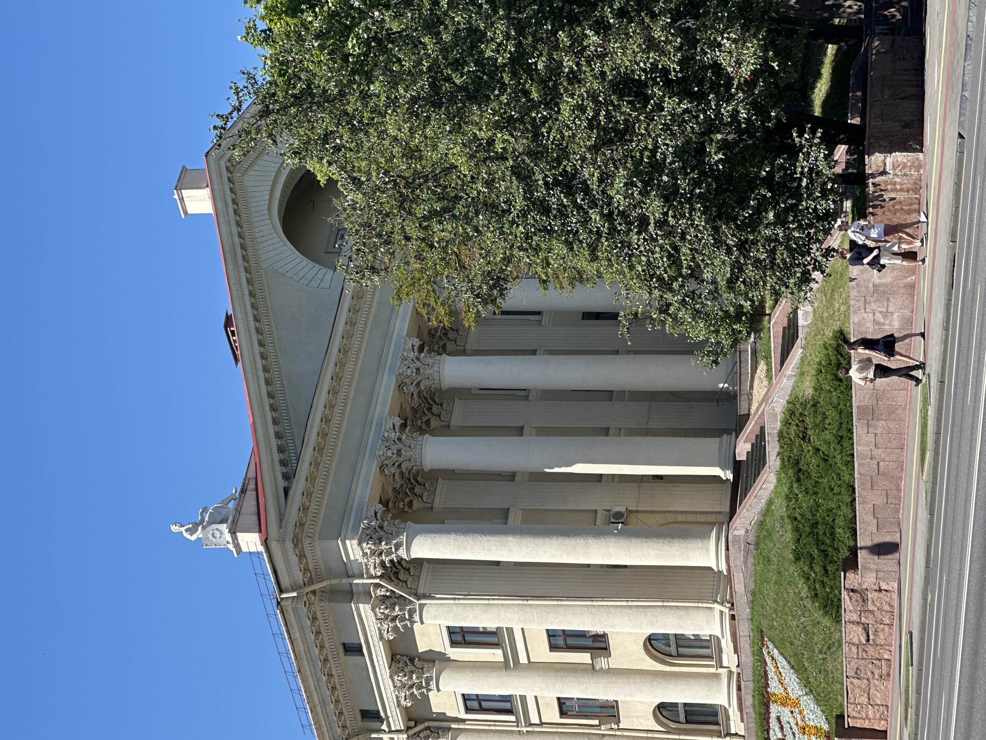 The image shows the exterior of the Ministry of Foreign Affairs of Belarus, located at 10 Lenin Street in Minsk, Belarus. The building features a neoclassical architectural style with a prominent colonnade of tall, fluted columns supporting an ornate entablature. A statue of a seated figure adorns the roofline above the central archway. In front of the building, a sloping, grassy lawn is decorated with flowerbeds. Several people are walking on the sidewalk in front of the building, on the left side of the image. A large, leafy tree partially obscures the right side of the building. The sky is clear and blue, indicating a sunny day. The scene is captured from a street-level perspective, with a road and sidewalk in the foreground.