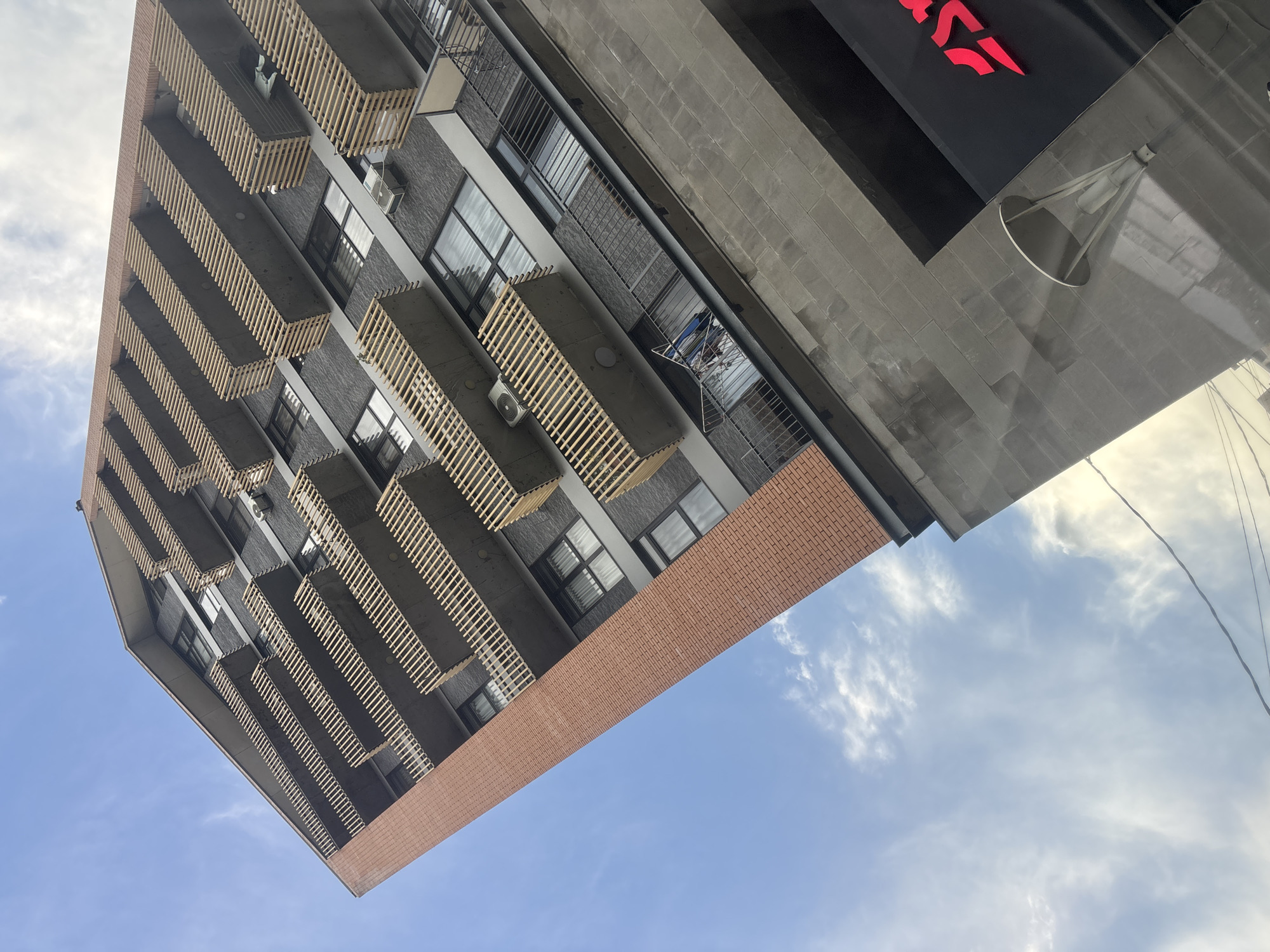 A low-angle shot shows the facade of a building against a blue sky with light clouds. The building has a modern design featuring a brick base and a gray upper section with multiple balconies. The balconies have light-colored wooden slats or screens, and air conditioning units are mounted beneath them. A portion of the building's upper right corner includes signage in an unreadable language, and an architectural element is visible. The location is likely Tbilisi, Georgia, based on the architectural style.