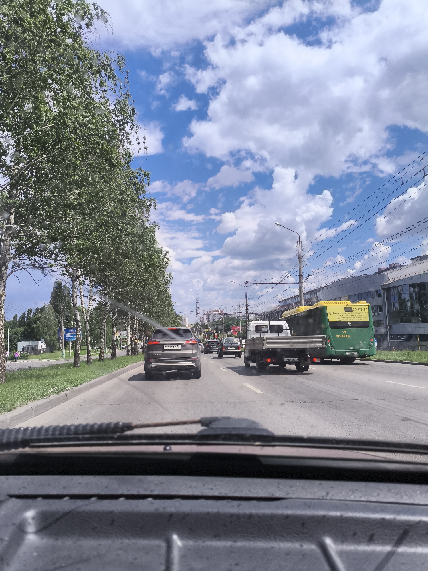 The image captures a view from inside a vehicle looking out at a street scene in Malaya Valyaevka, Russia. A line of birch trees with lush green foliage borders the right side of the road, which is paved with asphalt and marked with white lane lines. In the foreground, the dashboard and windshield of the vehicle are visible, with what appears to be water droplets or smudges on the glass.
Several vehicles are driving on the road. A dark-colored SUV is in the right lane, followed by a flatbed truck. Further ahead, a green and yellow bus is also in motion. The bus has writing on its back, including "ПЕНЗА" (Penza) and a date. In the distance, buildings and power lines can be seen. The sky is a vibrant blue, dotted with fluffy white clouds. A pedestrian is visible on the left side of the road, walking near a sign. The overall impression is a typical daytime scene on a suburban or urban street in Russia.