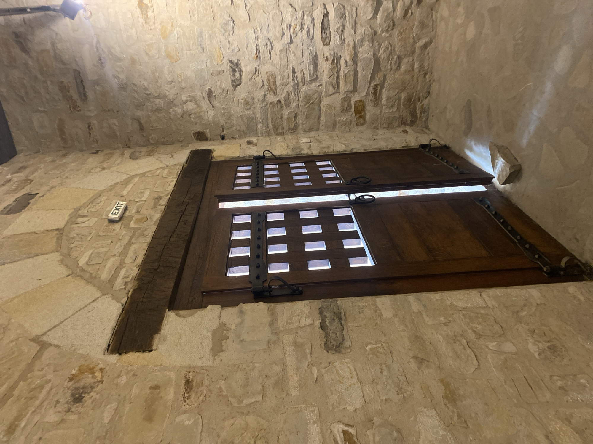 The image depicts an interior scene within a stone structure, possibly a historical building or fortification. A large, dark wooden double door with metal accents stands partially open, revealing a brightly lit space beyond. The door has a grid of small, square openings, allowing light to pass through. Above the doorway, a wooden beam is visible, supporting the stone archway. An illuminated "EXIT" sign is mounted on the stone wall to the left of the arch. The walls are constructed of rough, uneven stones, characteristic of older architecture. The floor is made of concrete or stone tiles. A small, light-colored stone is resting on the floor in front of the open doorway, illuminated by shafts of light. The environment appears to be dimly lit, with focused lighting from the "EXIT" sign and a ceiling-mounted fixture, parts of which are visible in the upper right corner. The location is identified as being within the city of Suceava, Romania.